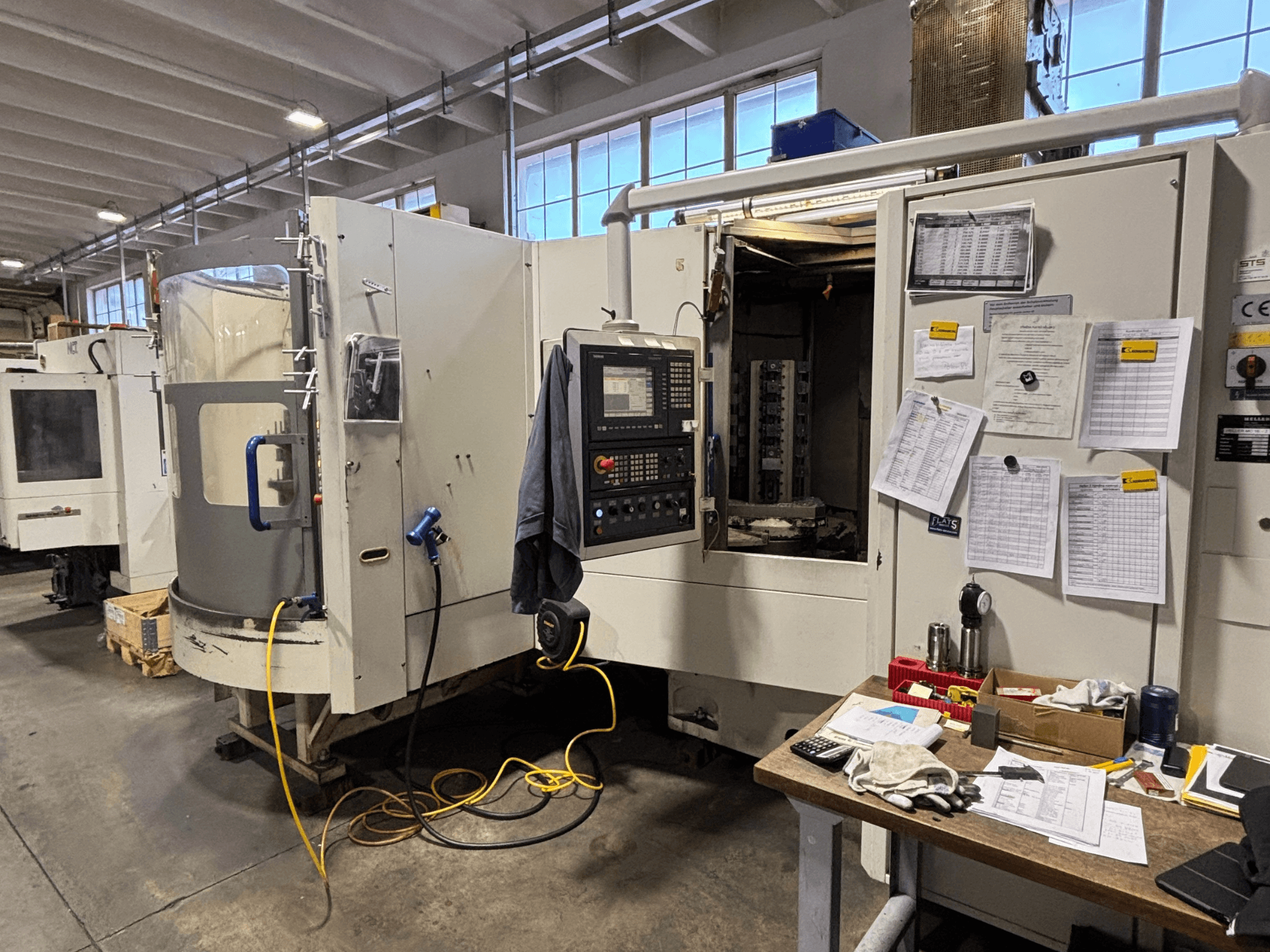 CNC stroj u radionici, prednji pogled, s upravljačkom pločom i dokumentacijom sa strane; alati i materijali na stolu.