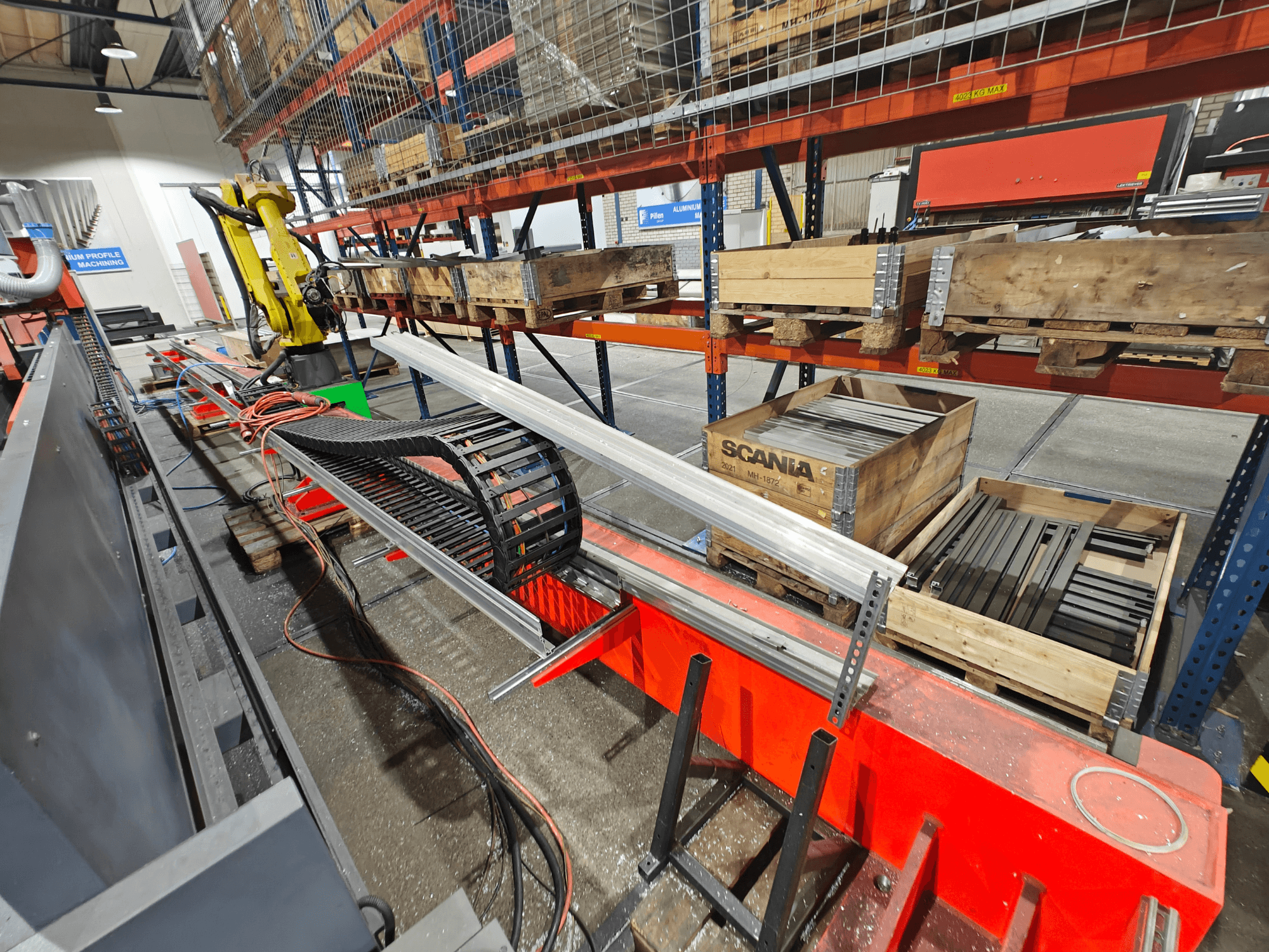 Robotska ruka na radnoj stanici, vidljivi narančasti i crni kabeli, sa Scania sanducima i aluminijskim profilima u tvorničkom okruženju.