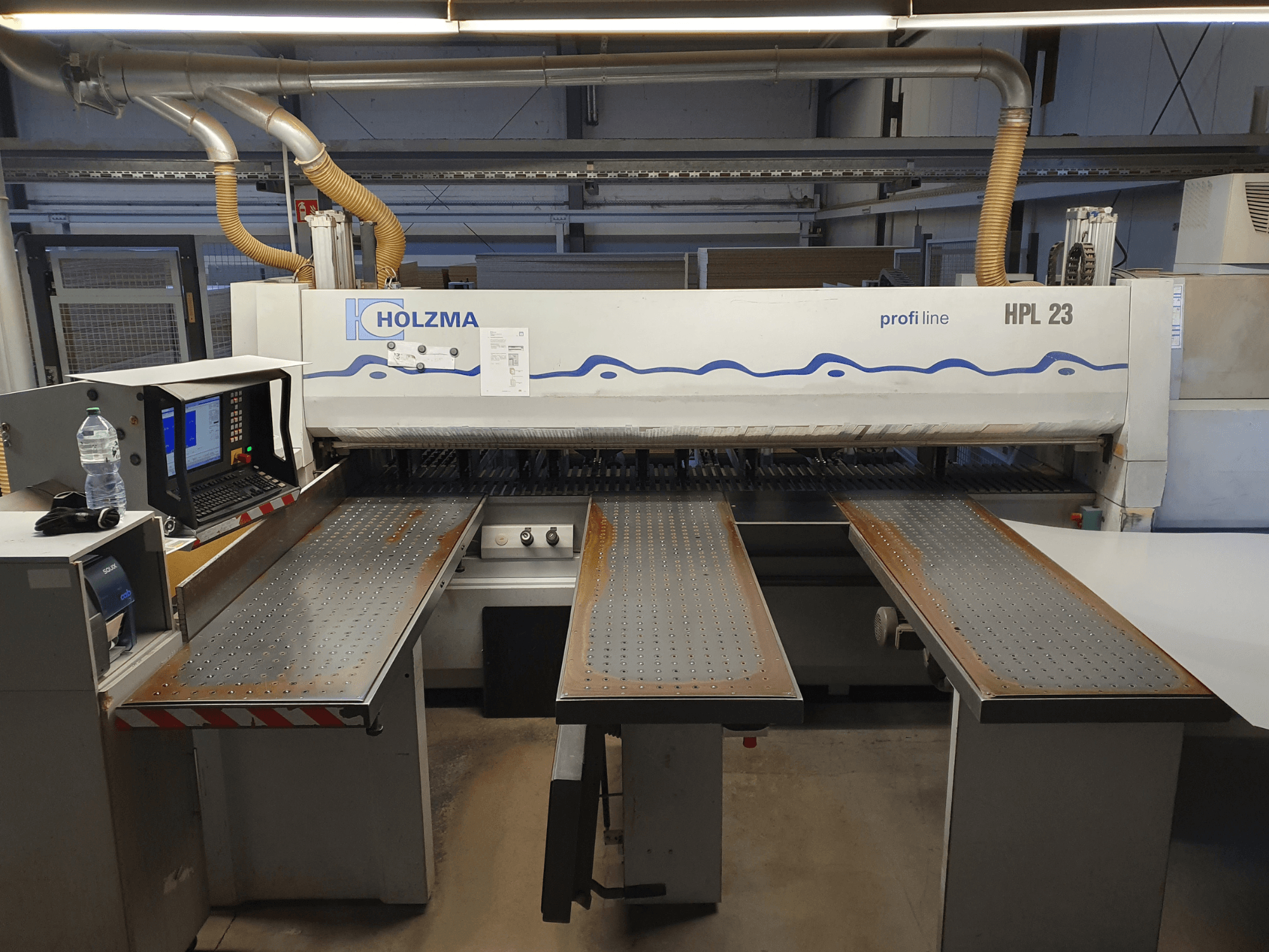 Stroj za rezanje HOLZMA HPL 23 prikazan s prednje strane, s tri perforirana stola, računalnim terminalom i bocom za vodu u blizini.