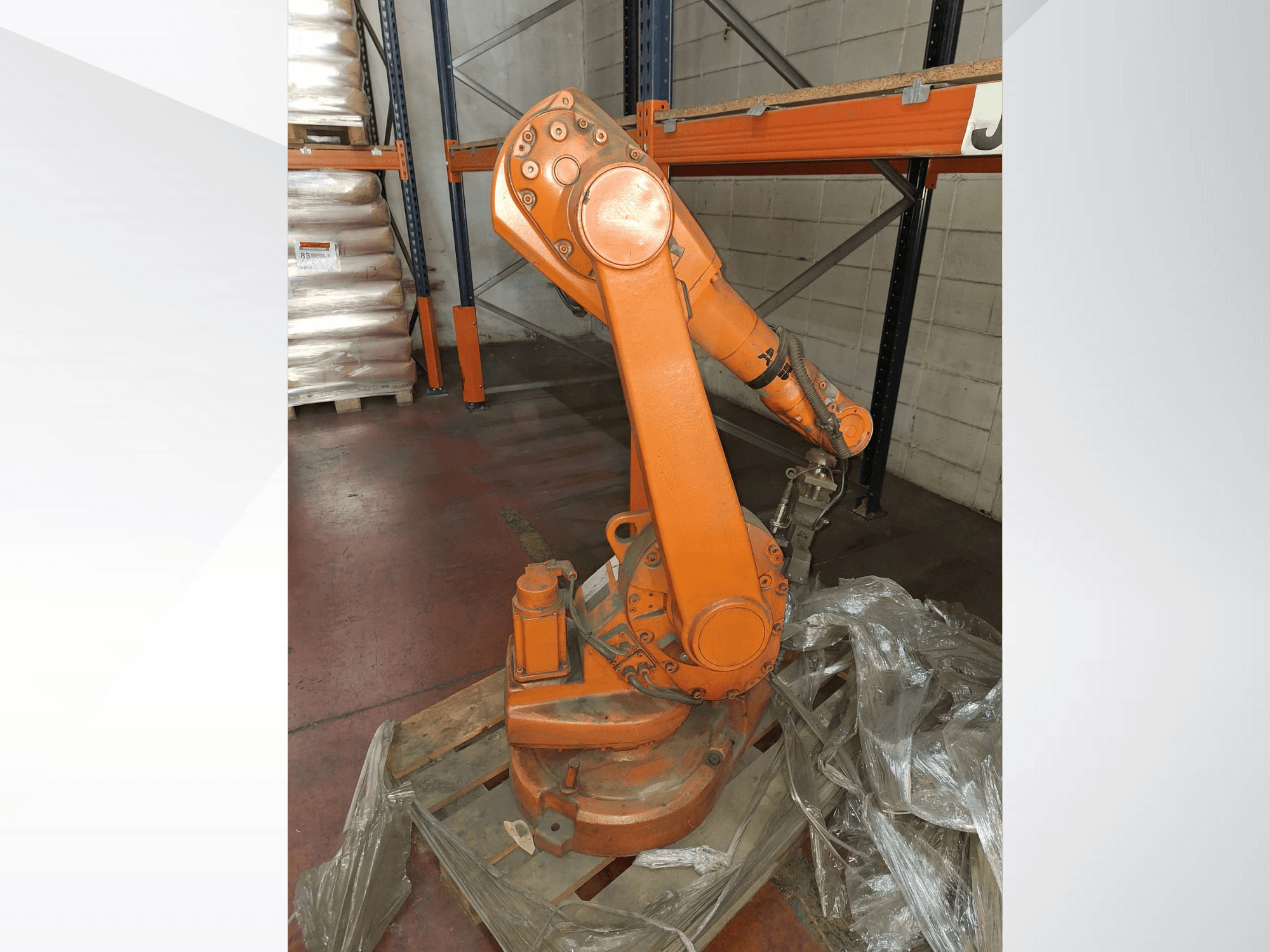 Industrijska robotska ruka u narančastoj boji, prednji pogled na drvenoj paleti, s pričvršćenim kabelom i zaštitnom plastičnom folijom oko baze.