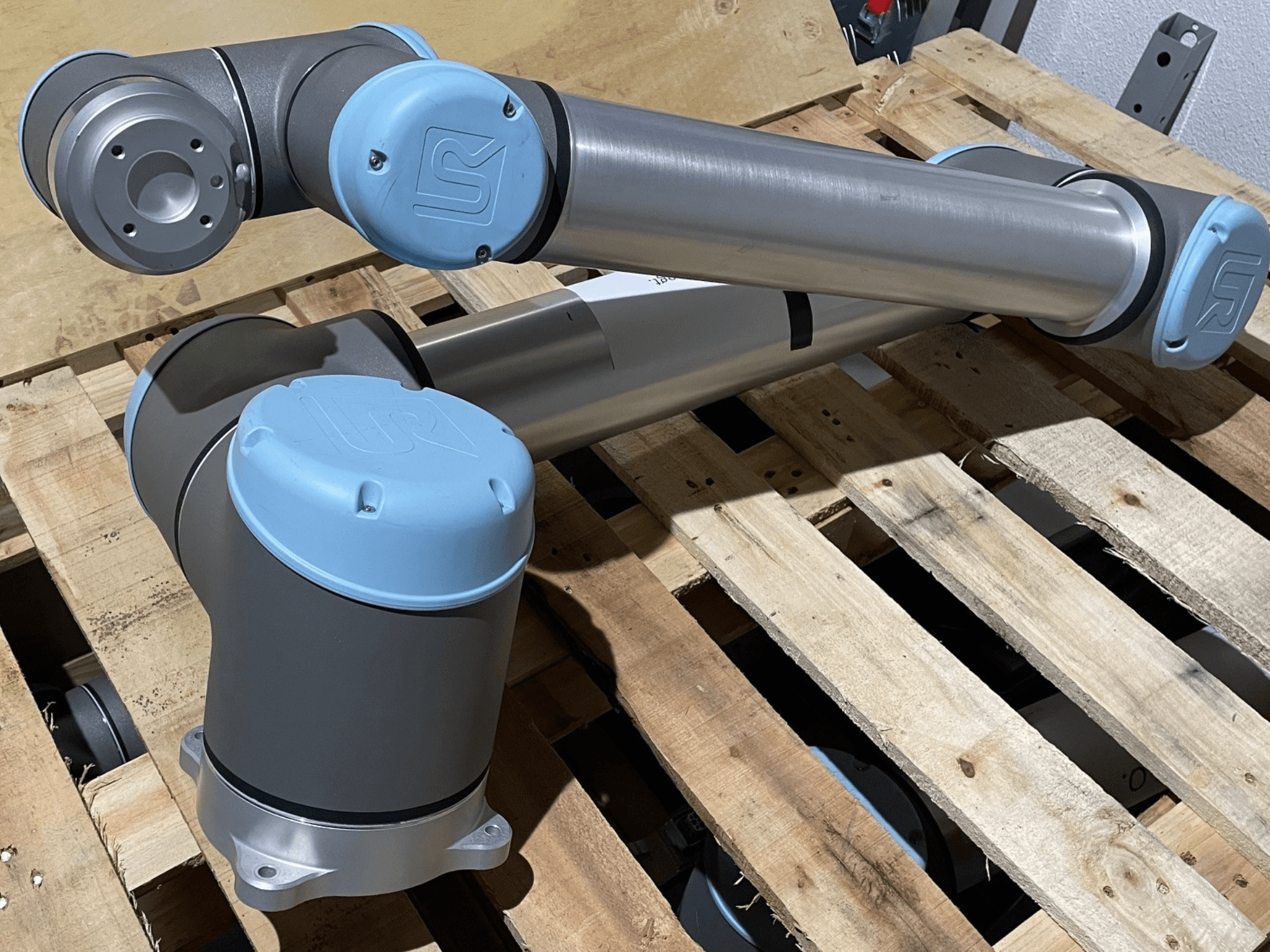 Srebrna robotska ruka s plavim završnim kapama, prednji pogled, prikazuje zglobove i mehaničke detalje, leži na drvenoj paleti.