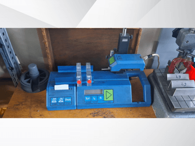 Plavi pisač Hommel T 1000 i tester T 1010 s crvenim gumbima, gledano sprijeda na radnom stolu s metalnim blokovima.
