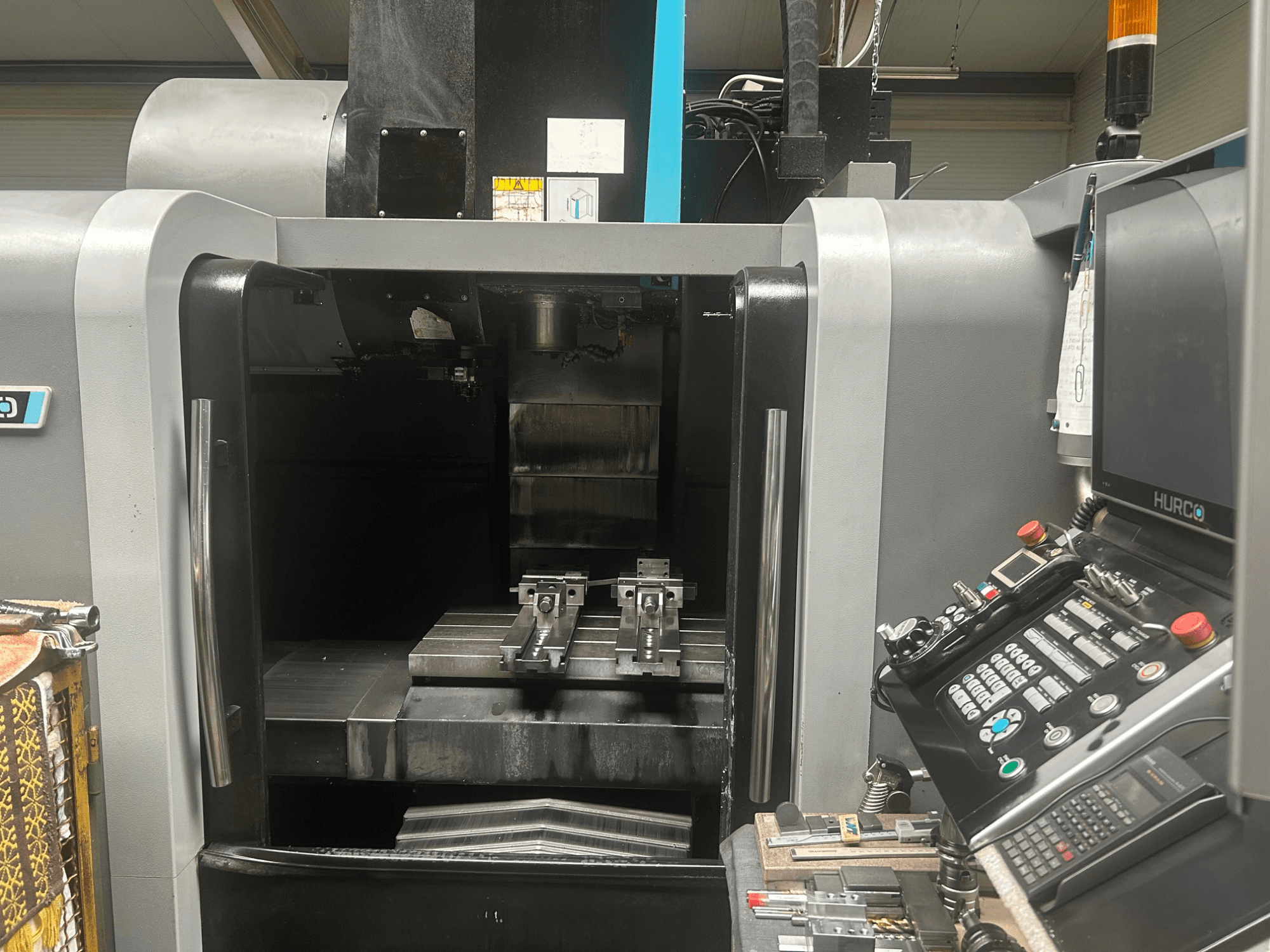 Prednji pogled na Hurco CNC obradni centar, koji prikazuje držač alata, metalni obratak i upravljačku ploču s gumbima i zaslonom.