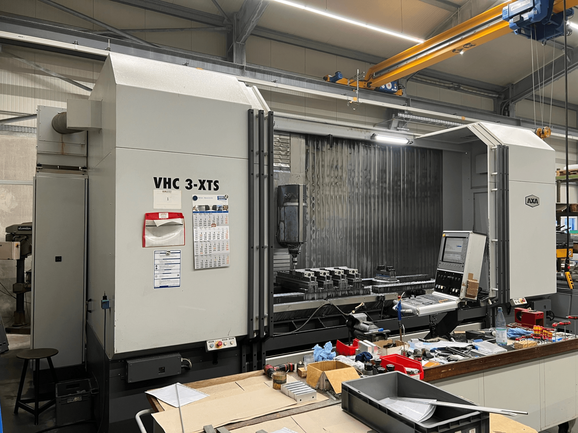AXA VHC 3-XTS CNC stroj u bočnom pogledu, s upravljačkom pločom i radnim prostorom zatrpanim alatima i komponentama.
