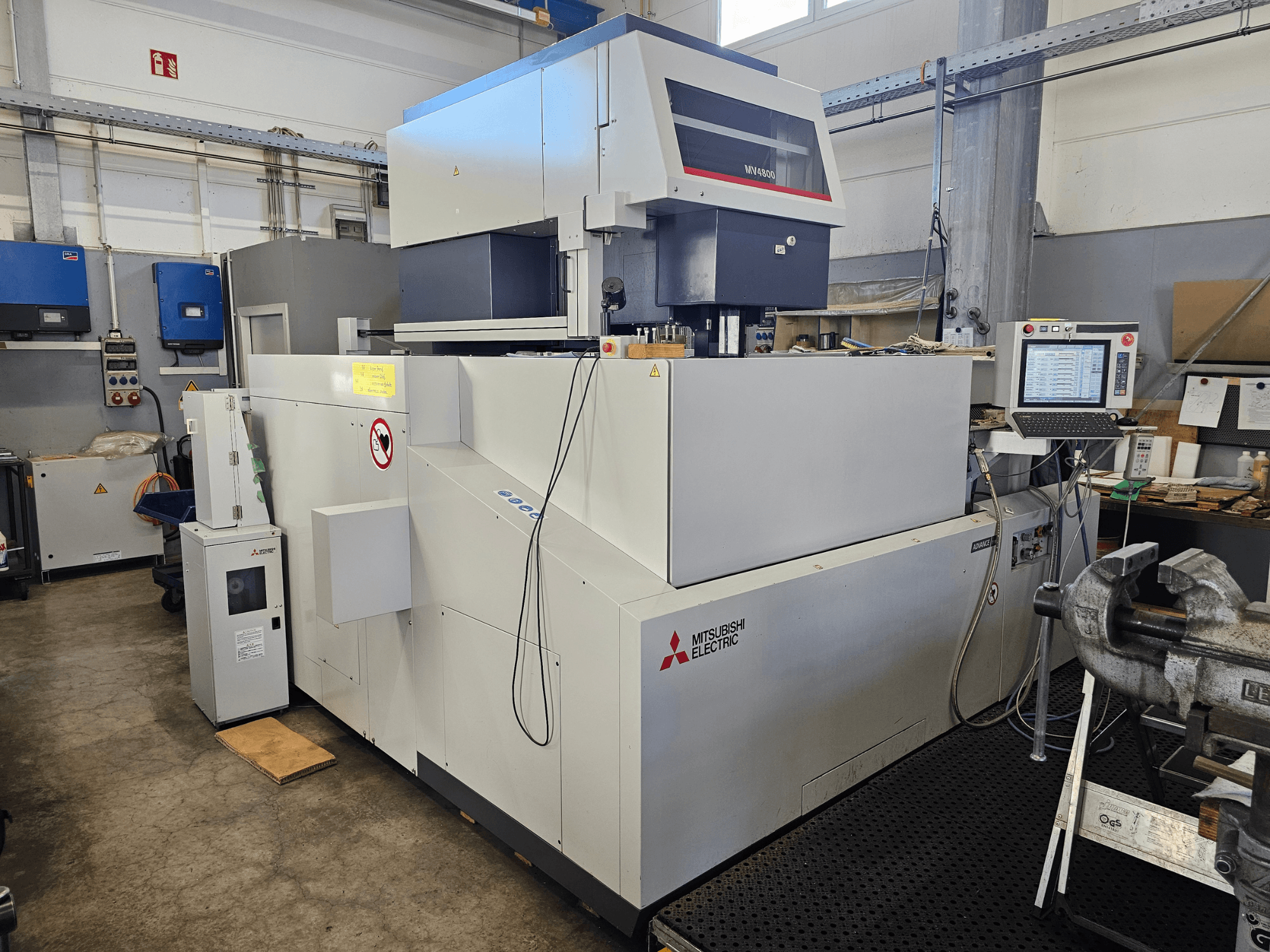 Stroj za žičanu eroziju Mitsubishi MV4800 u radionici, bočni pogled koji prikazuje upravljačku ploču, sigurnosne naljepnice i okolnu opremu.