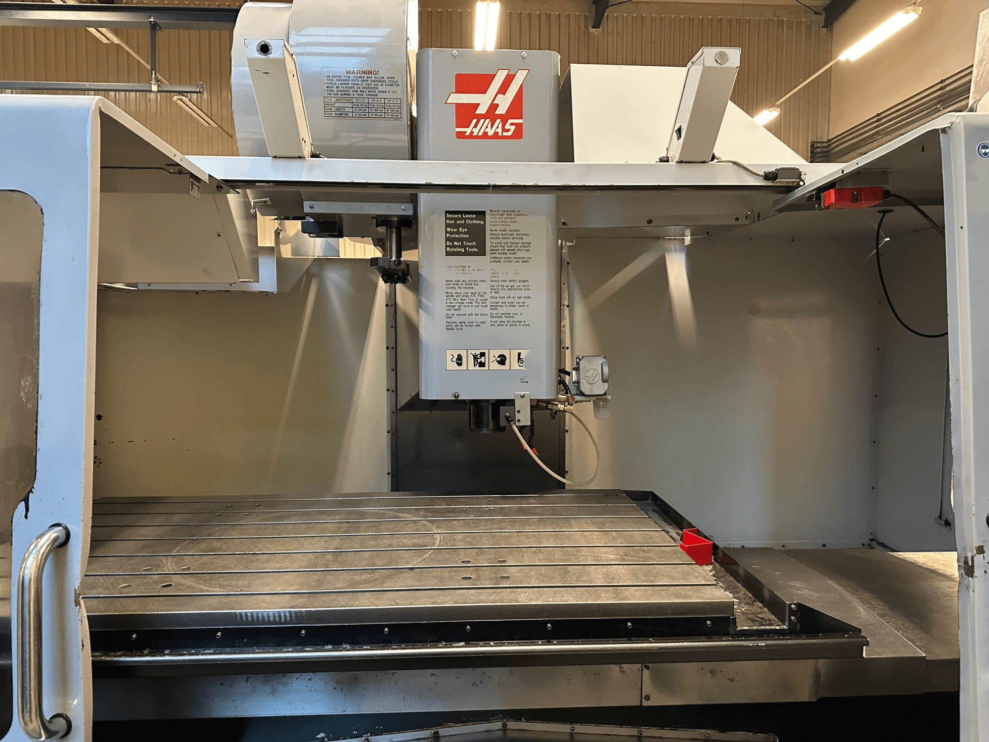 Haas CNC glodalica, model VF-2, prednji pogled s prikazom upravljačkih elemenata, metalnog radnog stola i sigurnosne naljepnice na ploči.