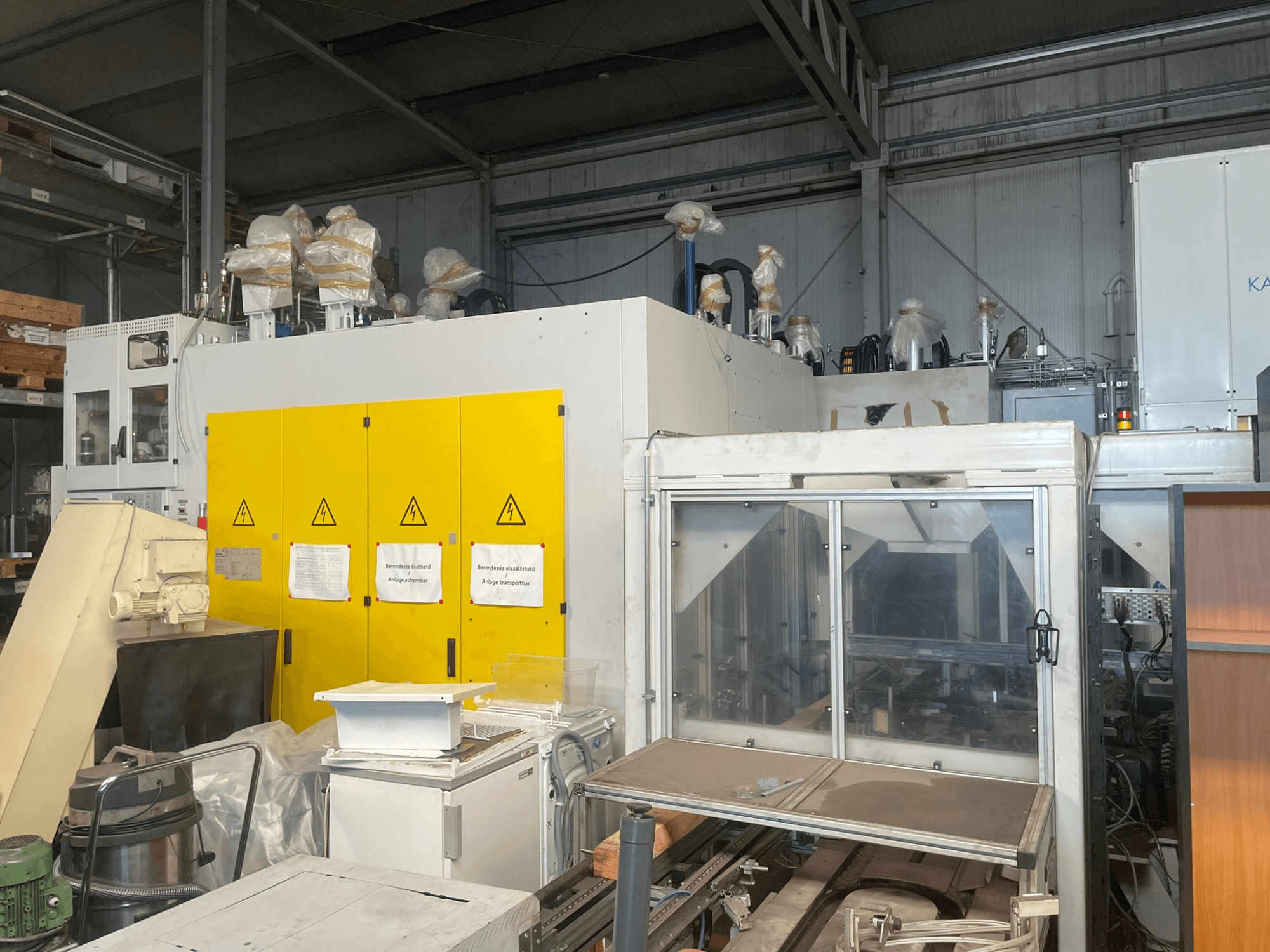 Industrijski strojevi sa žutom upravljačkom pločom, bočni pogled, s naljepnicama upozorenja i plastičnim poklopcima, u skladišnom okruženju.