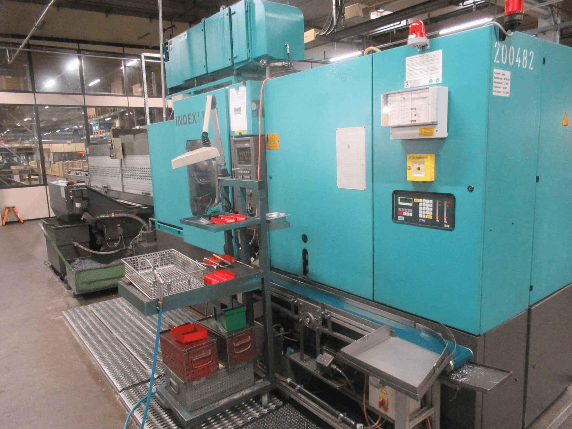 Tirkizni INDEX stroj u tvornici, bočni pogled; sadrži upravljačku ploču, spremište za alat i transporter. Vidljive kante, crijeva i alarmi.