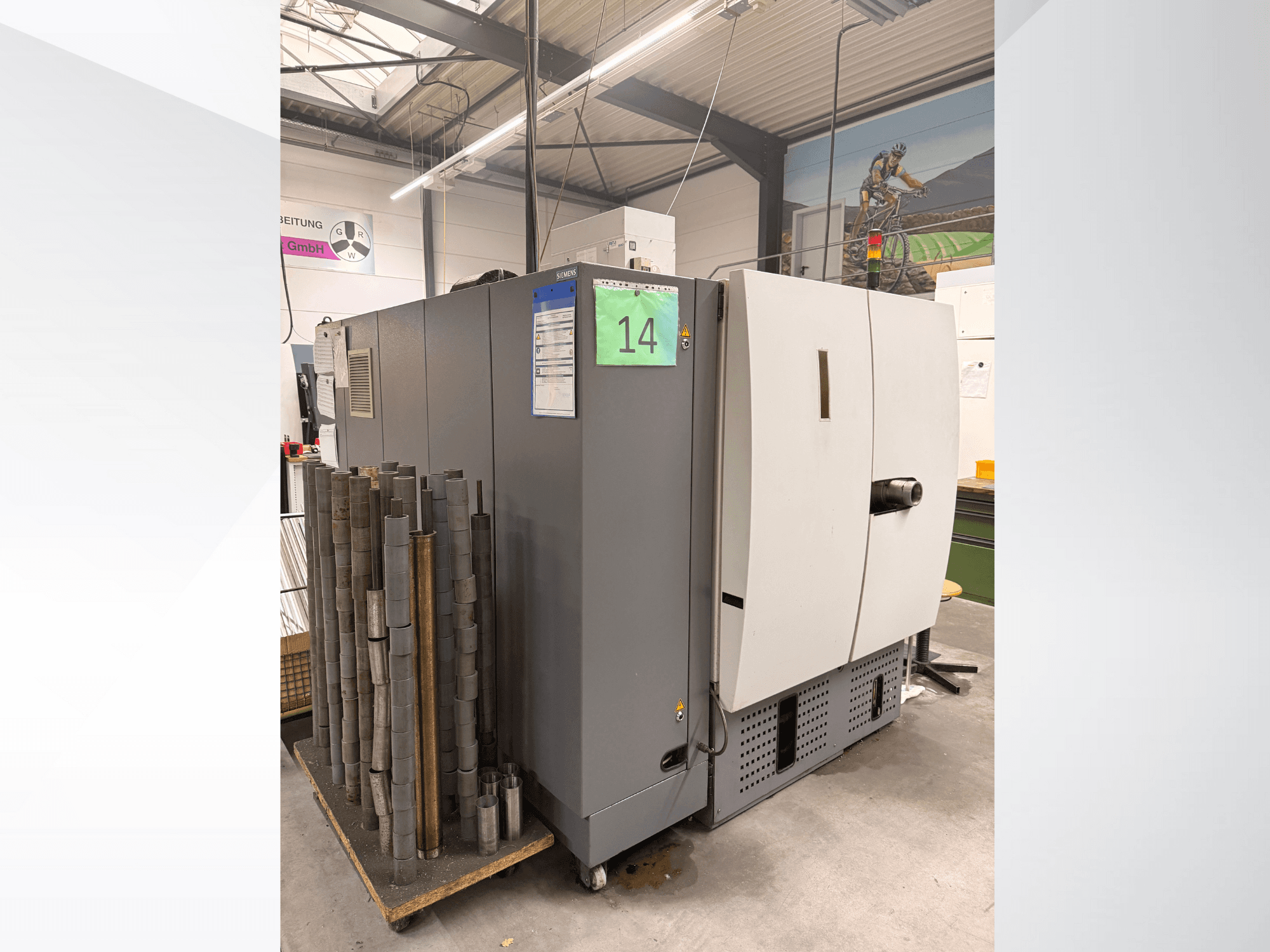 Sivi industrijski stroj tvrtke Siemens, bočni pogled, sa zalihom cilindričnih metalnih dijelova pored njega i vidljivim oznakama opreme.