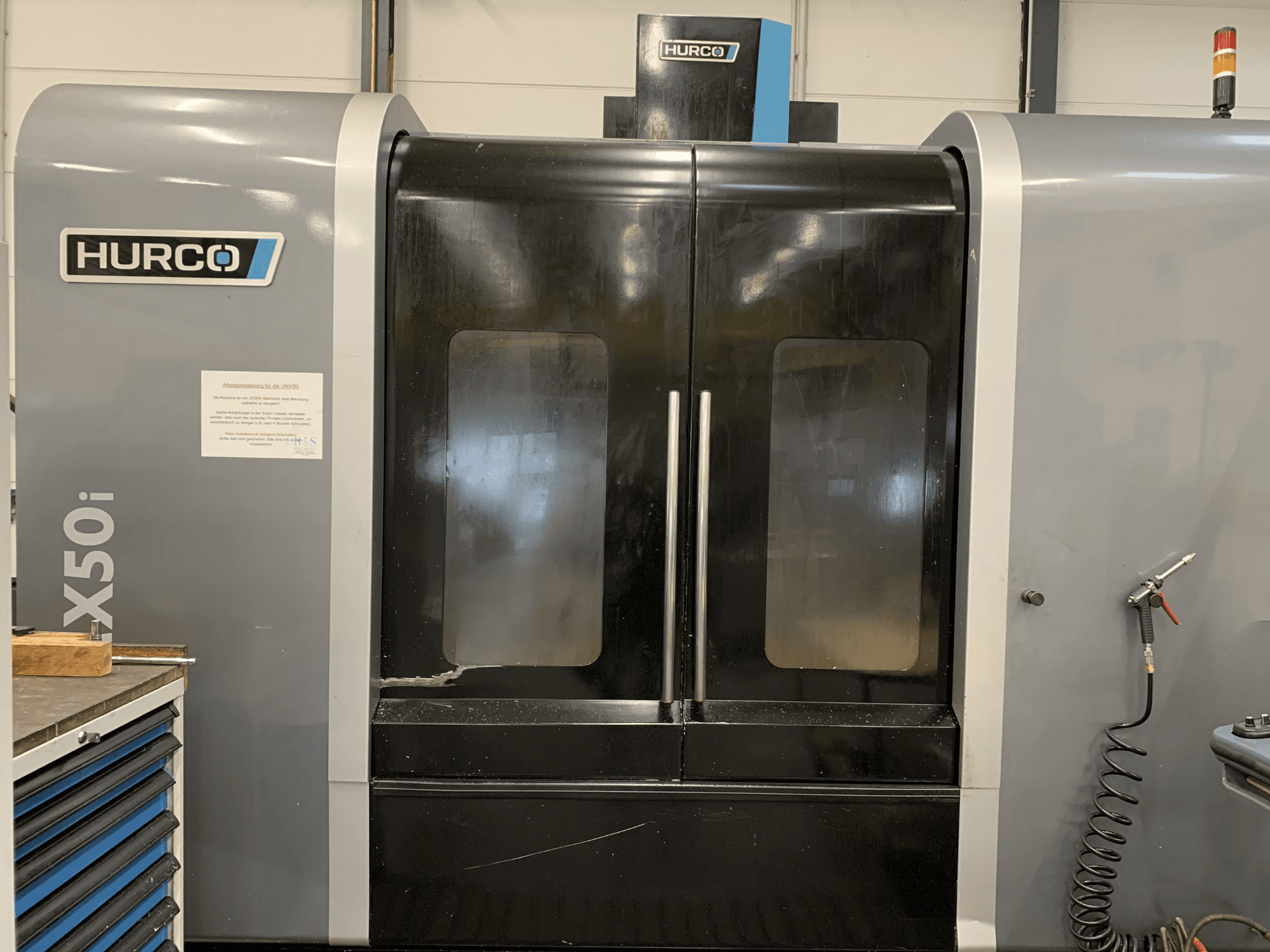 Prednji pogled na Hurco VX50i CNC stroj s dvostrukim vratima, s prikazom tehničkih kontrola i znaka za održavanje sa strane.
