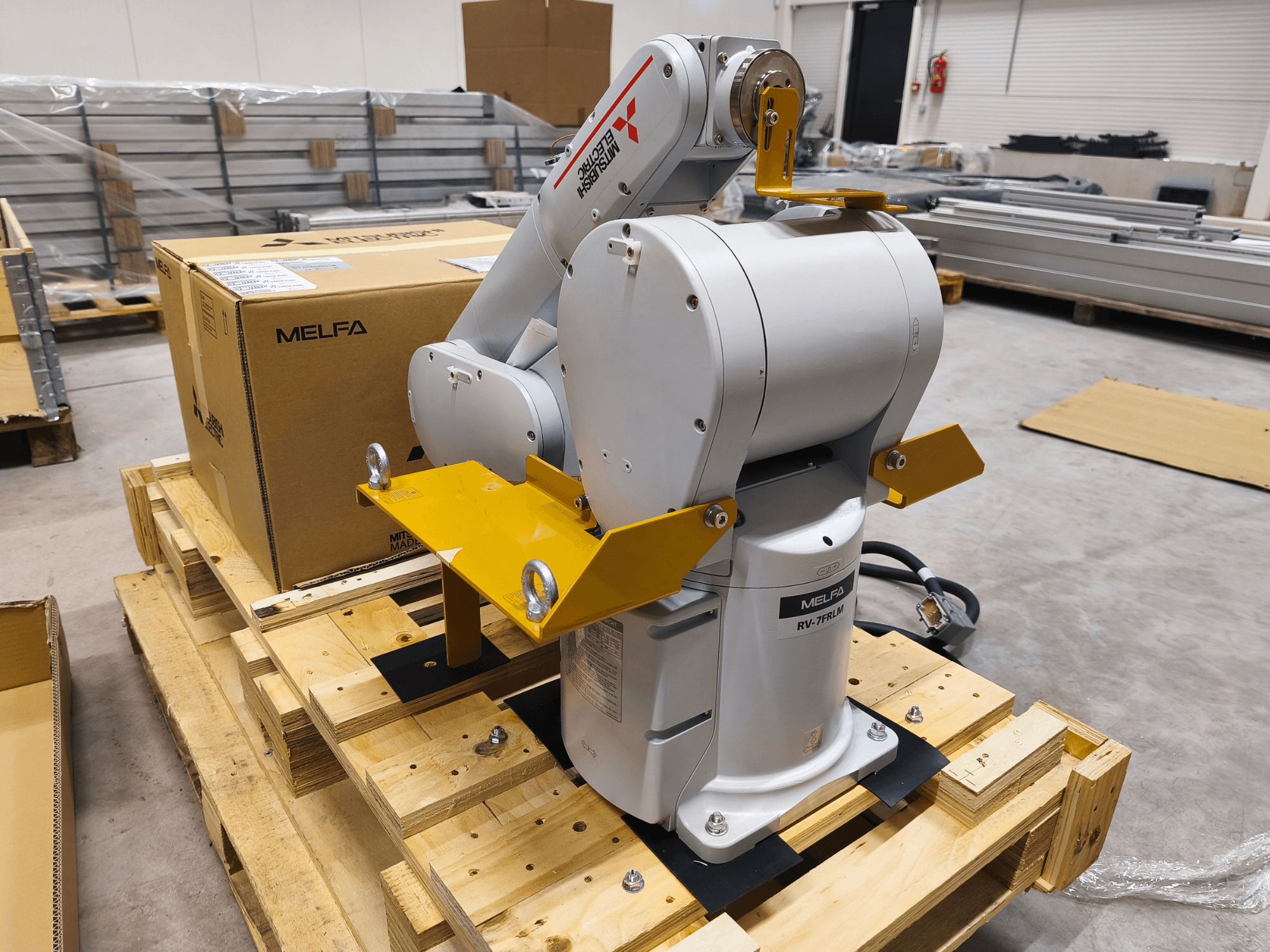 Industrijska robotska ruka MELFA RV-7FLM na drvenoj paleti, prednji pogled, sa žutim nosačima i sivom kutijom za pakiranje.