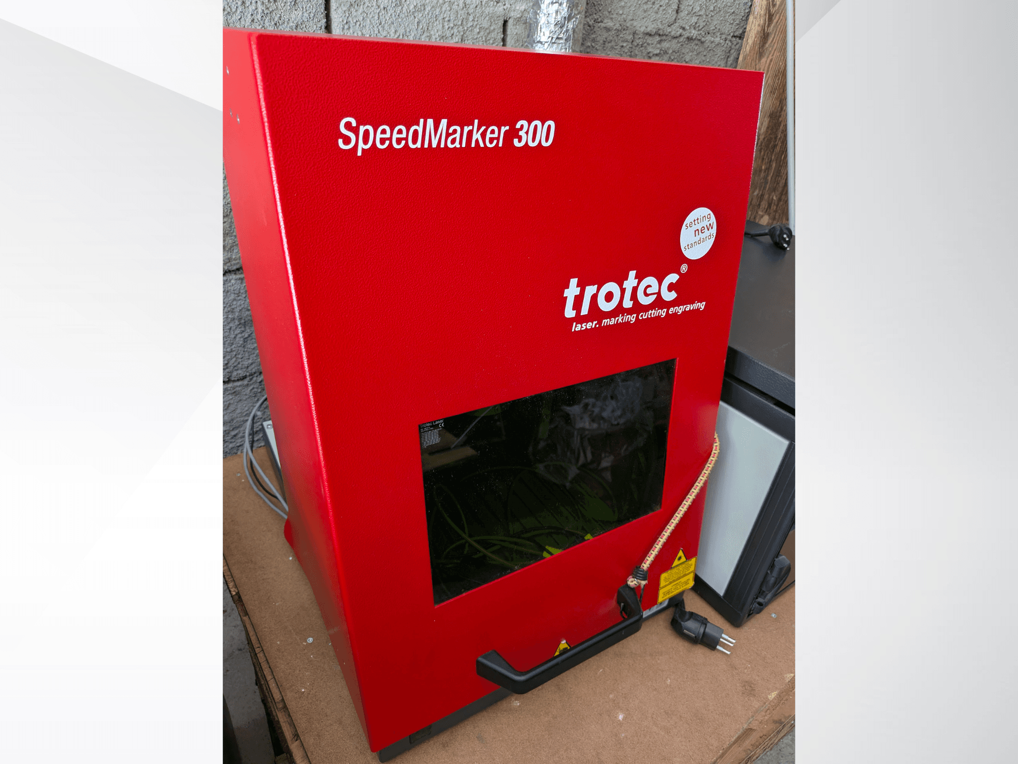 Crveni laserski stroj za označavanje Trotec SpeedMarker 300 u prednjem pogledu, s velikim prozorom za gledanje i priključkom za napajanje.