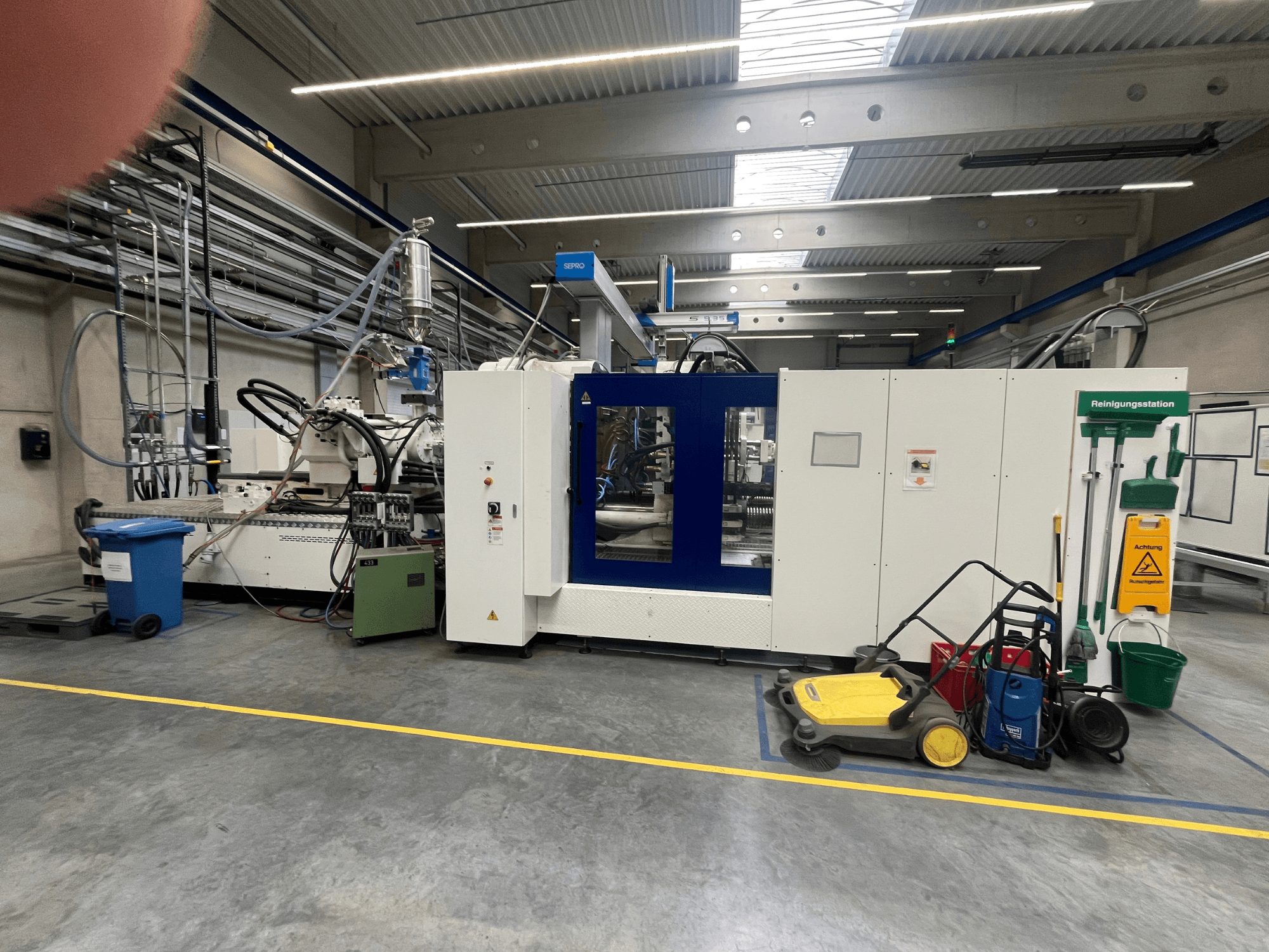 Industrijski stroj u tvornici, prednji pogled, s plavo-bijelim pločama, crijevima i opremom za čišćenje u blizini.