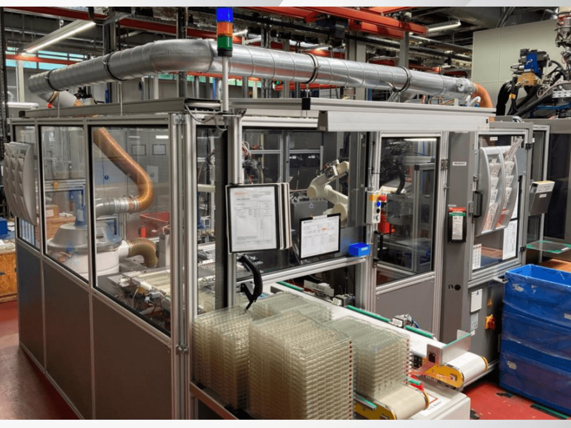 Prednji pogled na automatizirani stroj s metalnim cijevima, upravljačkom pločom i naslaganim prozirnim plastičnim spremnicima na tvorničkom podu.