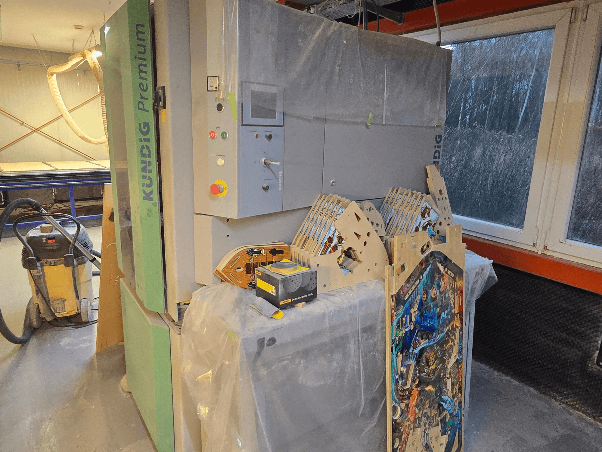 Tračna brusilica Kundig Premium-1 1100-C u radionici, s prikazom upravljačke ploče i drvenih dijelova, bočni pogled, s usisavačem prašine u pozadini.