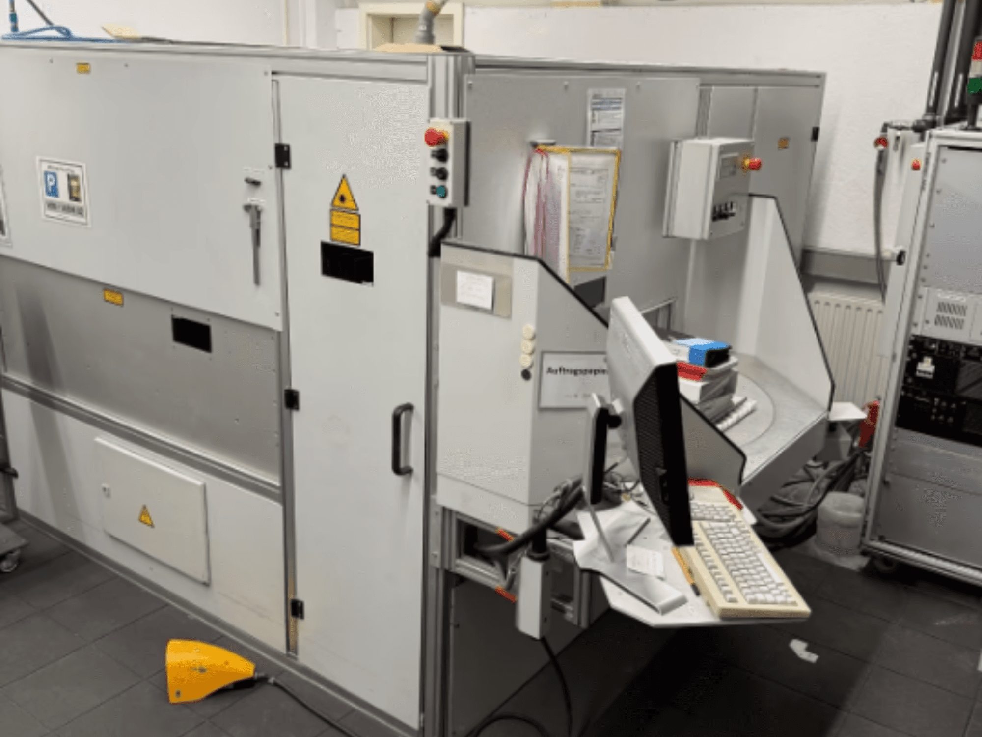 Industrijski stroj s računalnim terminalom, bočni pogled; uključuje upravljačke tipke i sigurnosne znakove upozorenja.