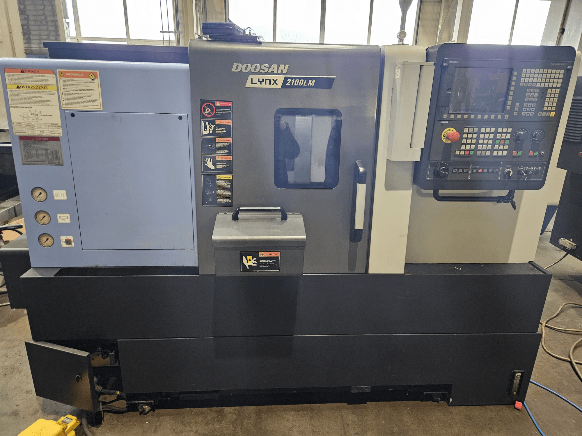 CNC tokarski stroj Doosan Lynx 2100LM, prednji pogled, s vidljivim upravljačkom pločom, mjeračima tlaka zraka i sigurnosnim naljepnicama.