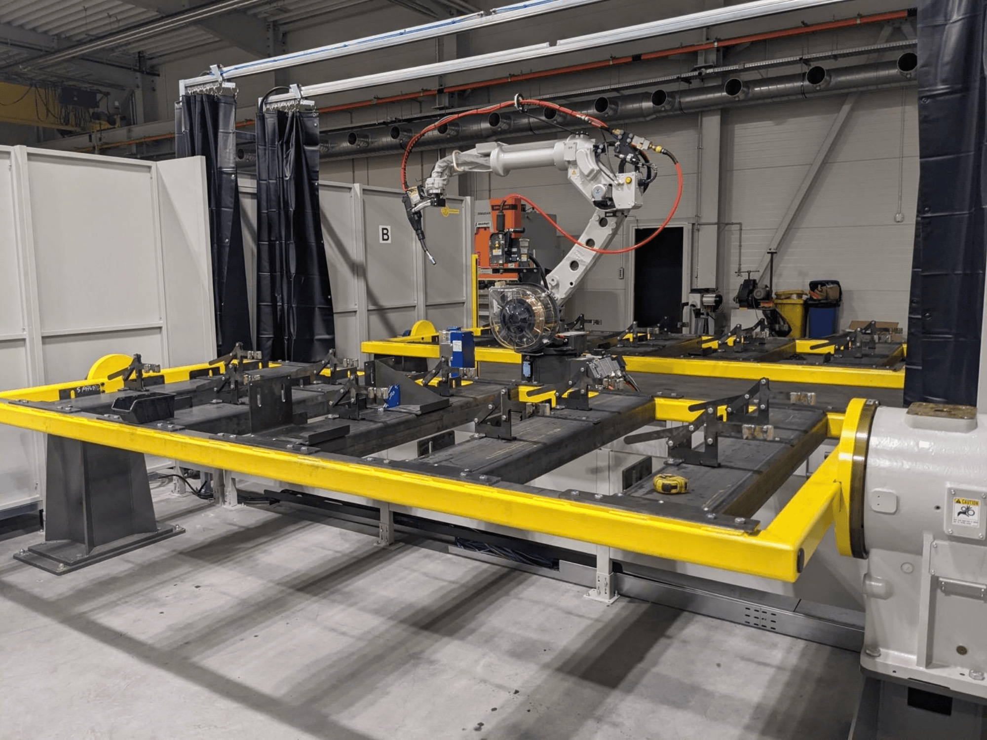 Prednji pogled na robotsku ruku PANASONIC TAWERS TM-2000WG3 postavljenu iznad žutog radnog stola s metalnim dijelovima za montažu.