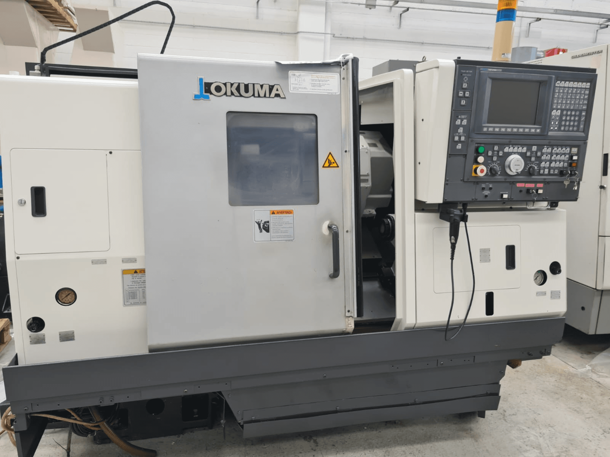 Horizontalni tokarski stroj Okuma LB 300M X 500 u bočnom pogledu, s prikazom upravljačke ploče, pristupa vretenu i sigurnosnih upozorenja.