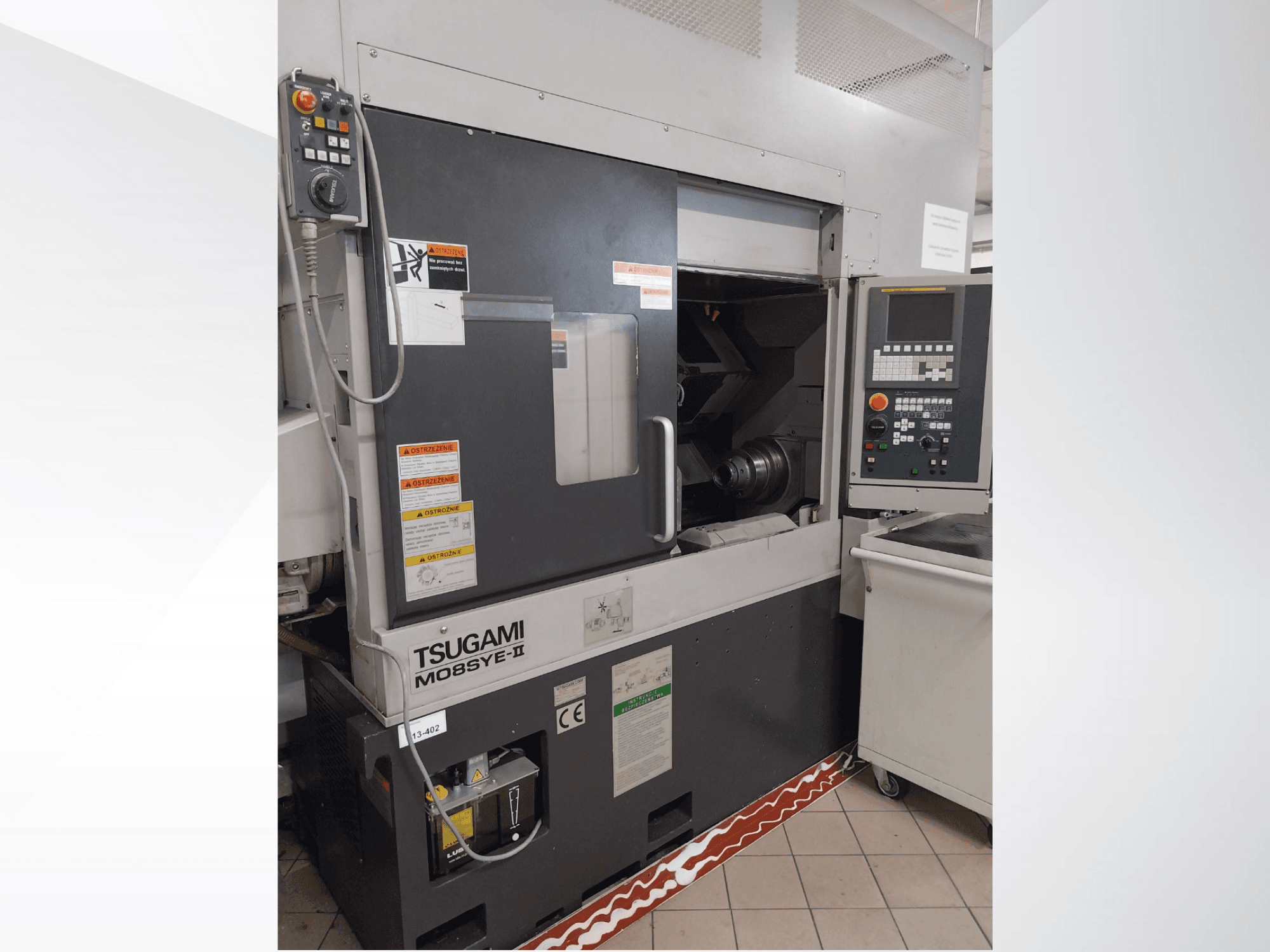 Tsugami M08SY-II CNC tokarski stroj, prednji pogled, prikazuje upravljačku ploču, vreteno i područje za utovar. Sadrži sigurnosne napomene i naljepnicu robne marke.