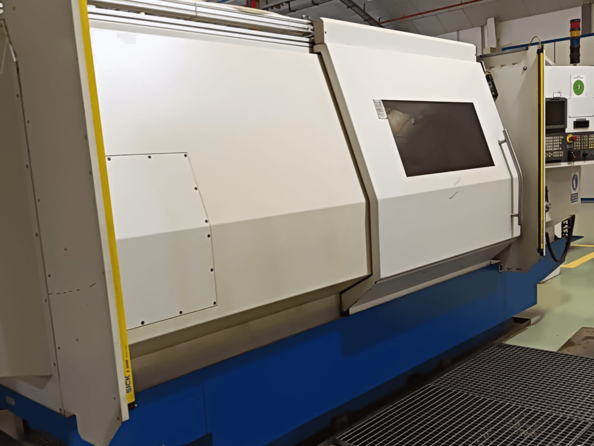 Industrijski tokarski stroj, bočni pogled, s velikim bijelo-plavim kućištem, digitalnom upravljačkom pločom i sigurnosnim značajkama.