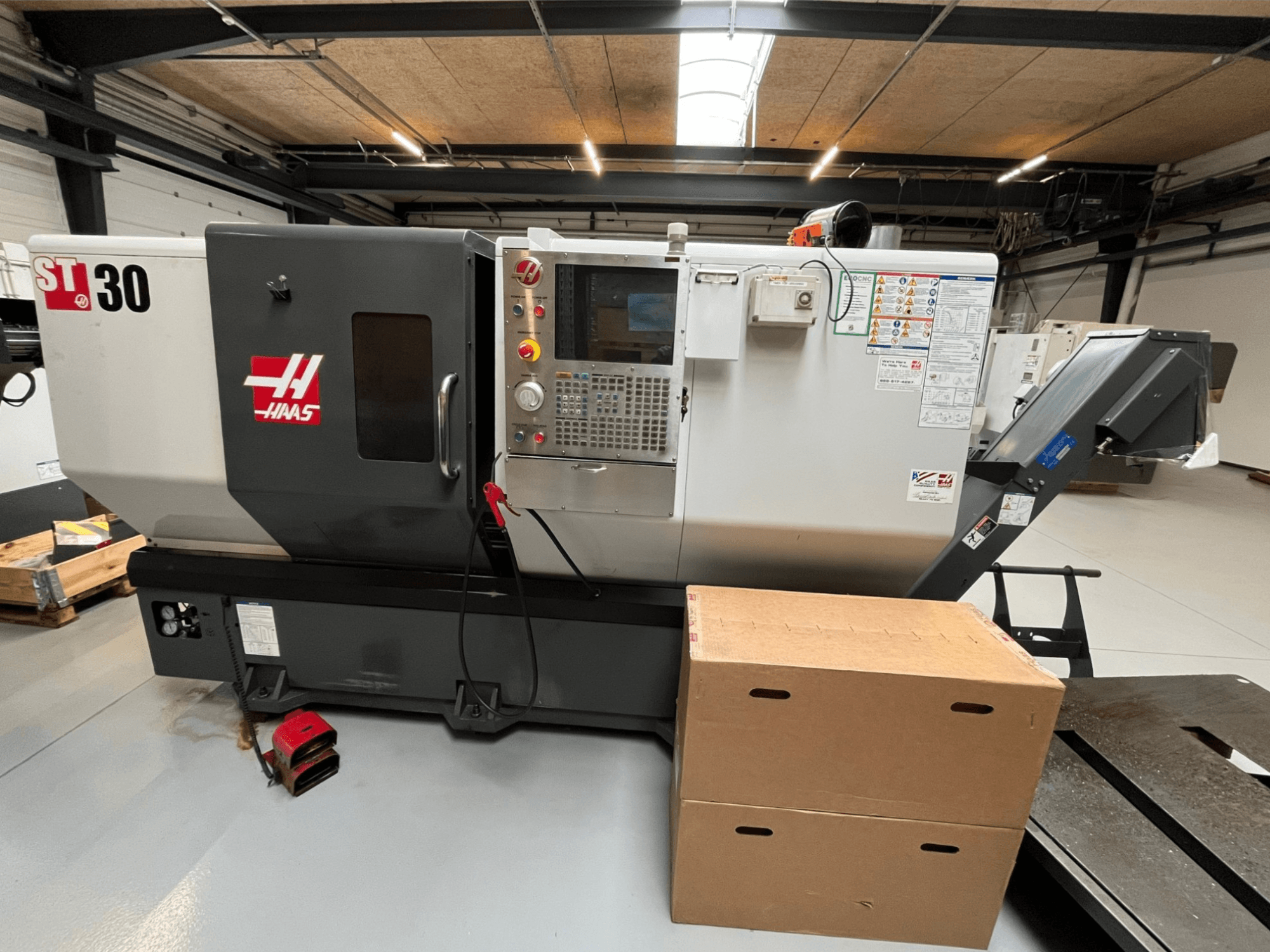 Haas ST-30 CNC tokarski stroj, prednji pogled, ima upravljačku ploču s gumbima, zaustavljanje u nuždi i dvije naslagane kartonske kutije.