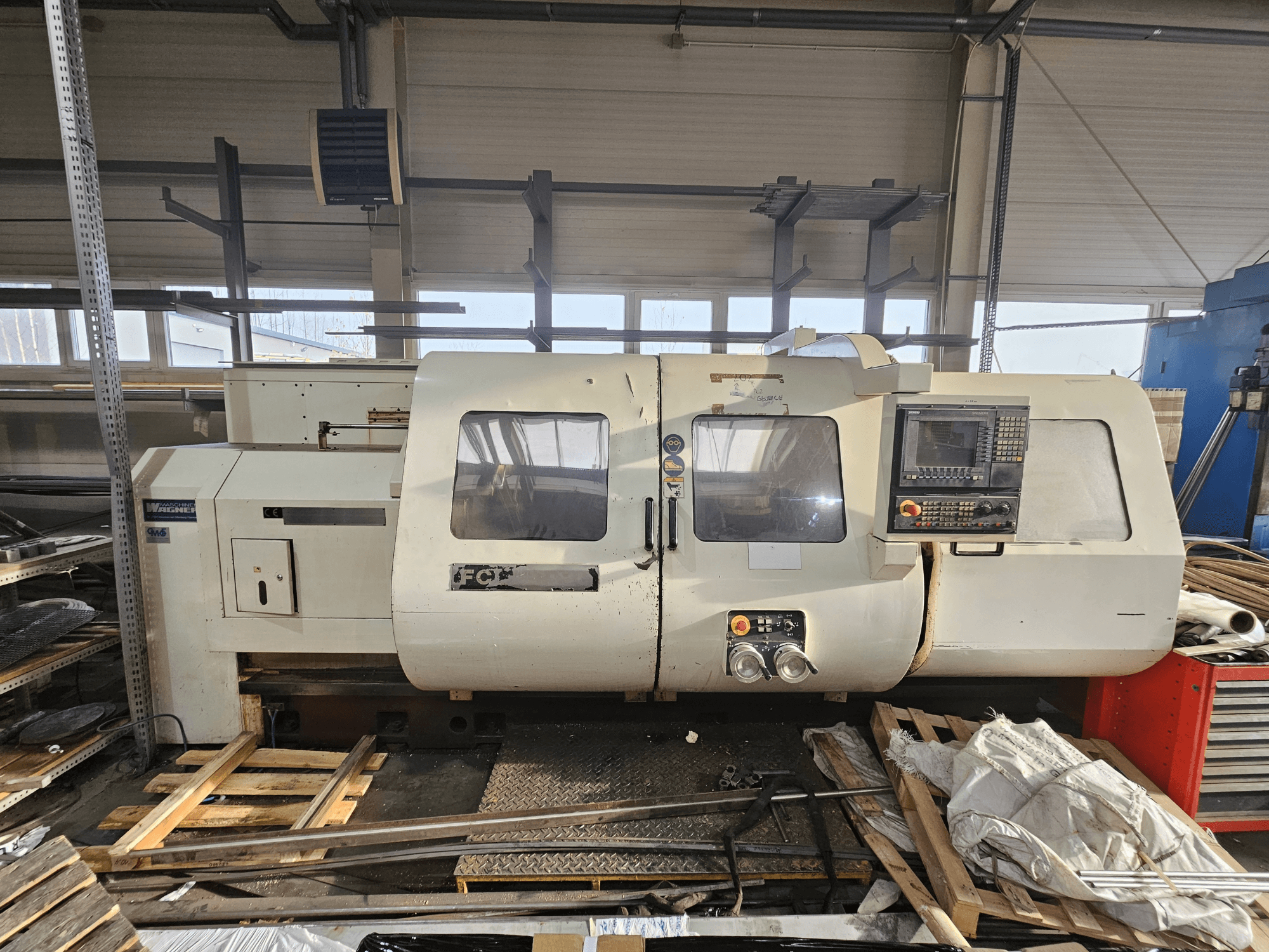 Wabeco CNC tokarski stroj, bočni pogled, s upravljačkom pločom, prozorima i radnim prostorom; strojevi na drvenim paletama u radioničkom okruženju.