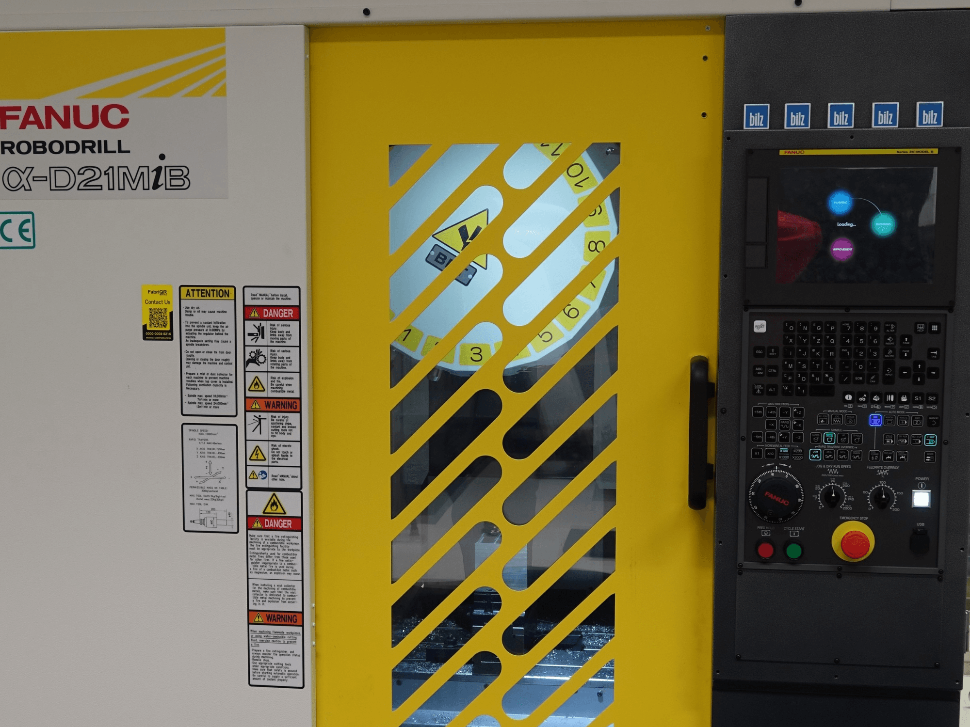 Prednji pogled na vertikalni obradni centar FANUC α-D21MiB, s prikazom sigurnosnih upozorenja, upravljačke ploče i žutih sigurnosnih vrata.