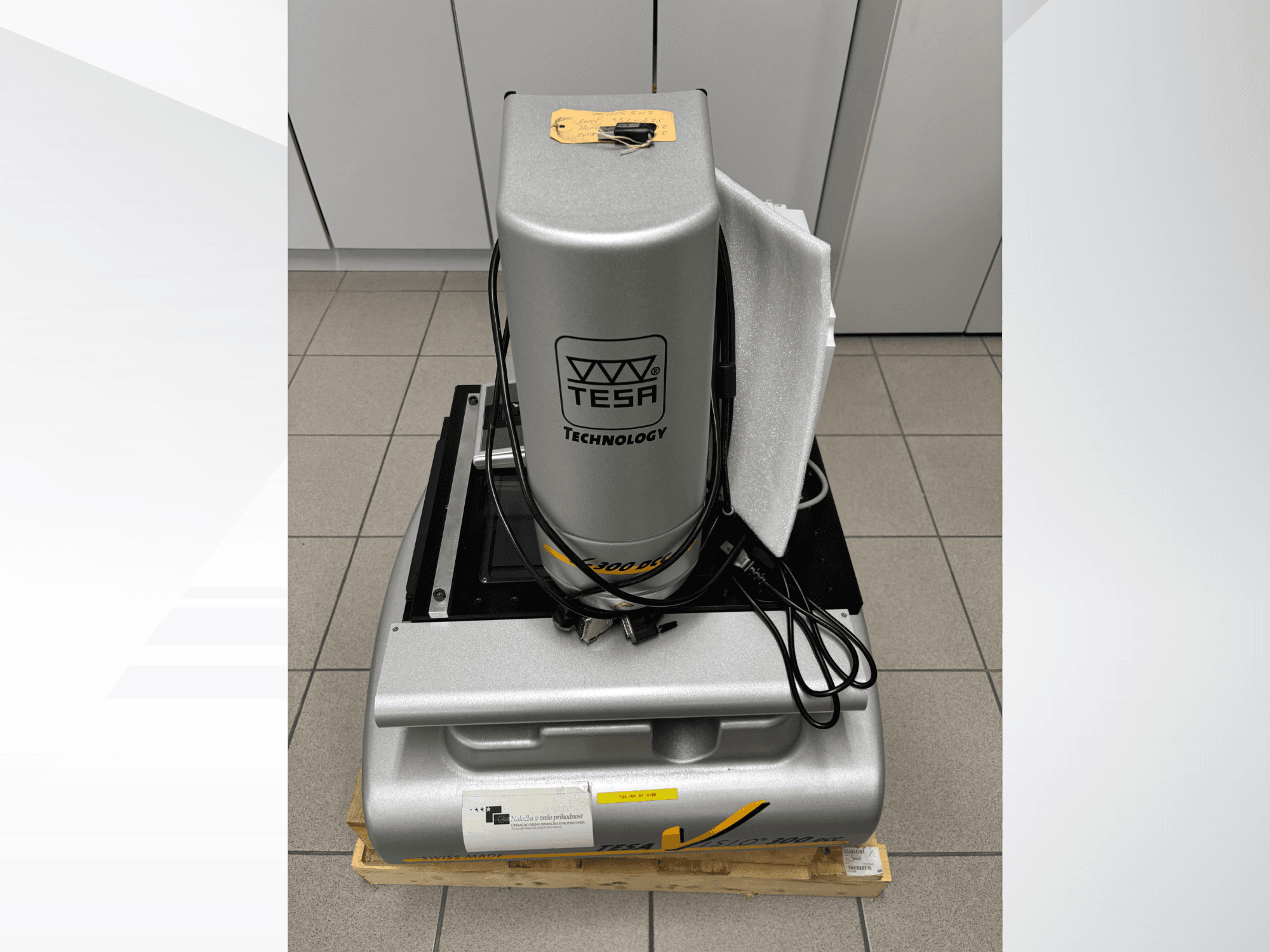 Optički mjerni stroj TESA Visio 300 DCC u prednjem pogledu s kabelima, na drvenoj paleti i vidljivom zaštitnom ambalažom.