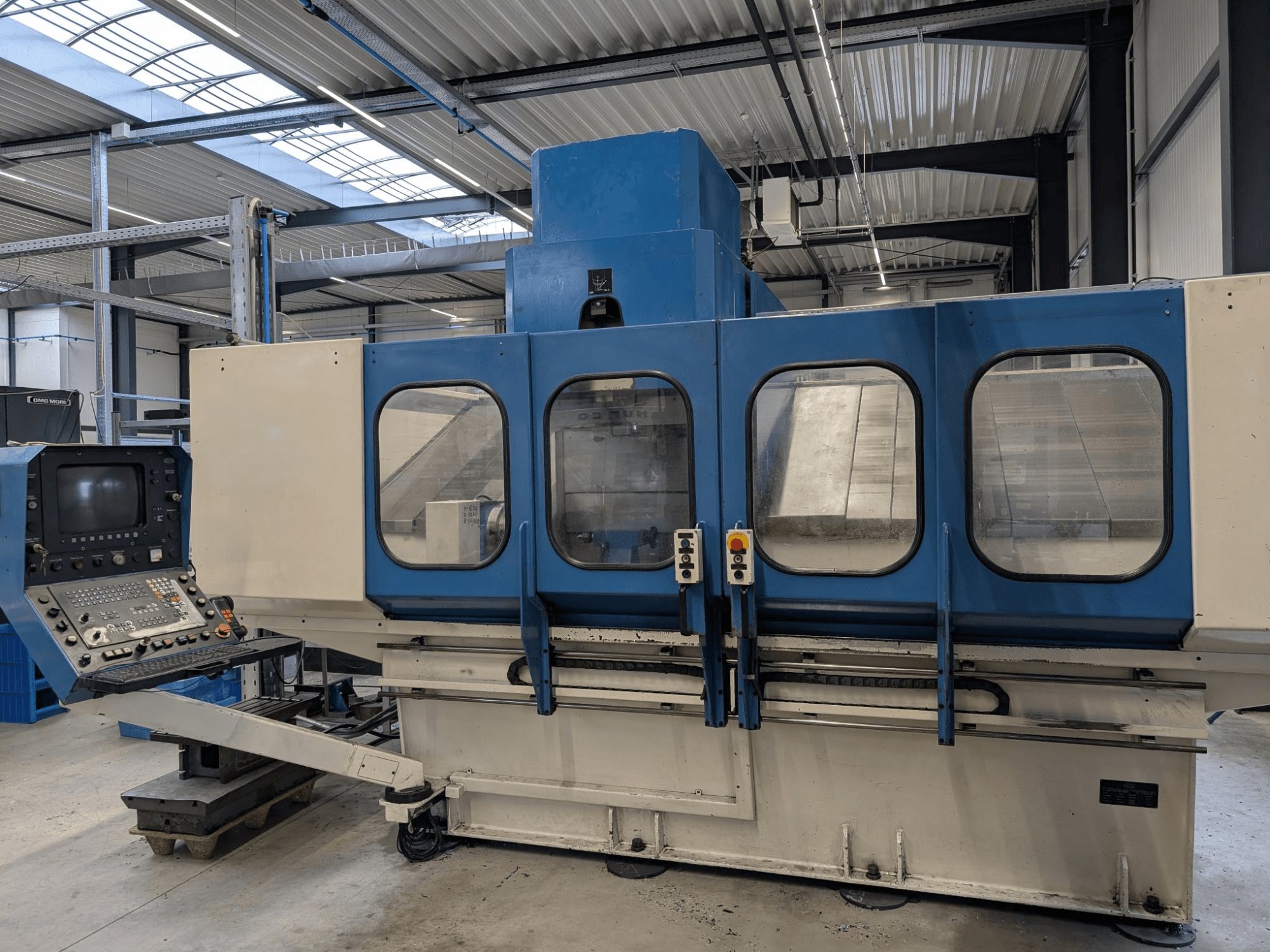 Plavi industrijski CNC stroj s upravljačkom pločom, prednji pogled. Veliki stakleni prozori, vidljive strojne komponente i gumbi.
