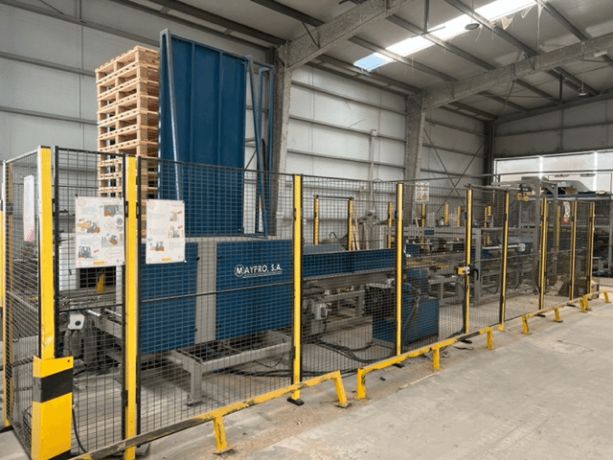 Industrijski stroj za pakiranje paleta tvrtke Maypro SA u skladištu, sa sigurnosnom mrežom i operativnim kontrolama, bočni pogled.