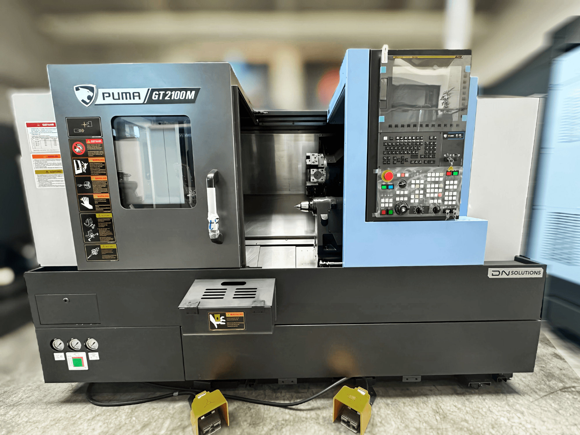 Tokarsko-glodački centar DN SOLUTIONS PUMA GT2100M, prednji pogled s upravljačkom pločom, komorom za obradu i sigurnosnim naljepnicama.