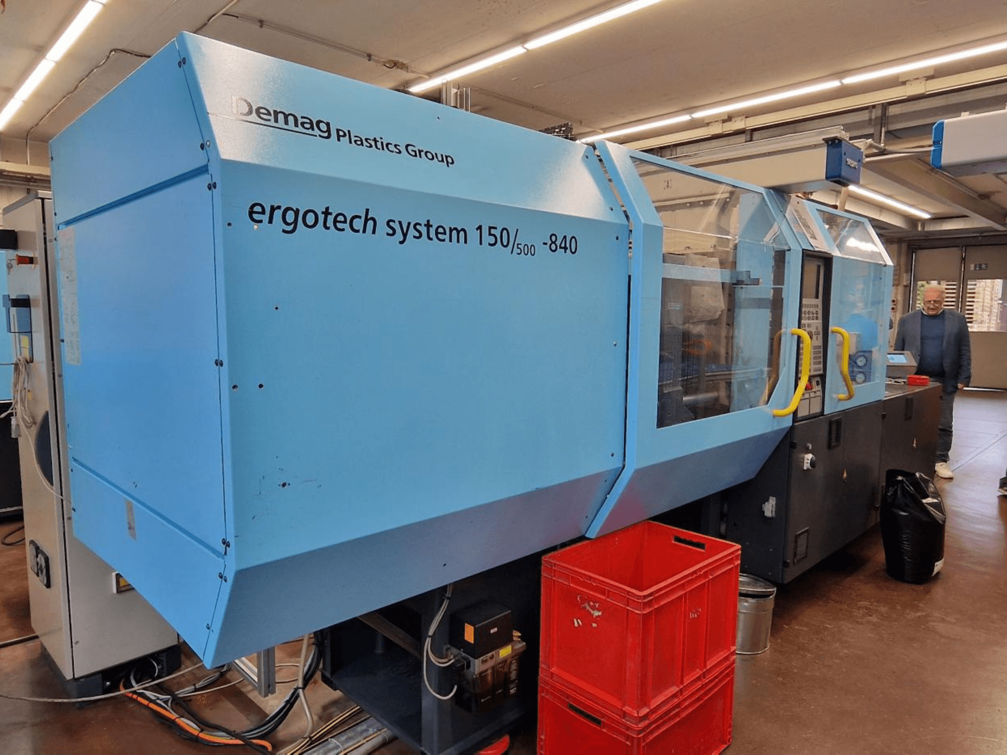 Stroj za brizganje plastike Demag Ergotech System 150/500-840 u plavoj boji, bočni pogled, s kutijama i operaterom u pozadini.