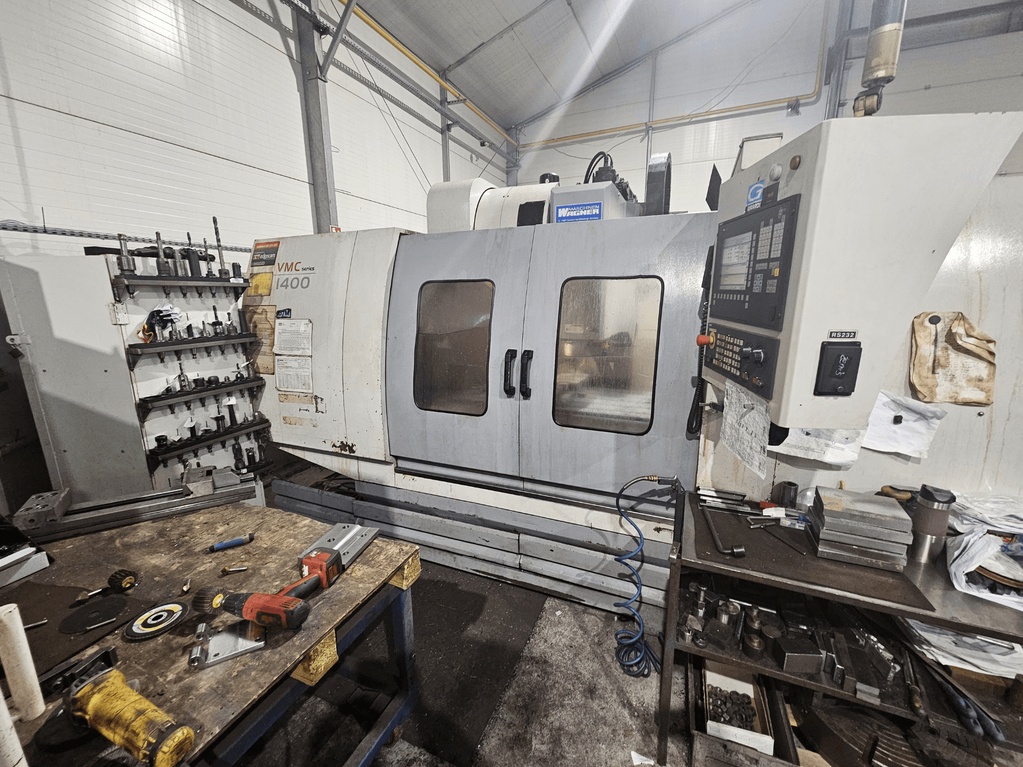 CNC glodalica VMC 1400 u radionici, prednji pogled; alati i materijali razbacani po obližnjem radnom stolu.