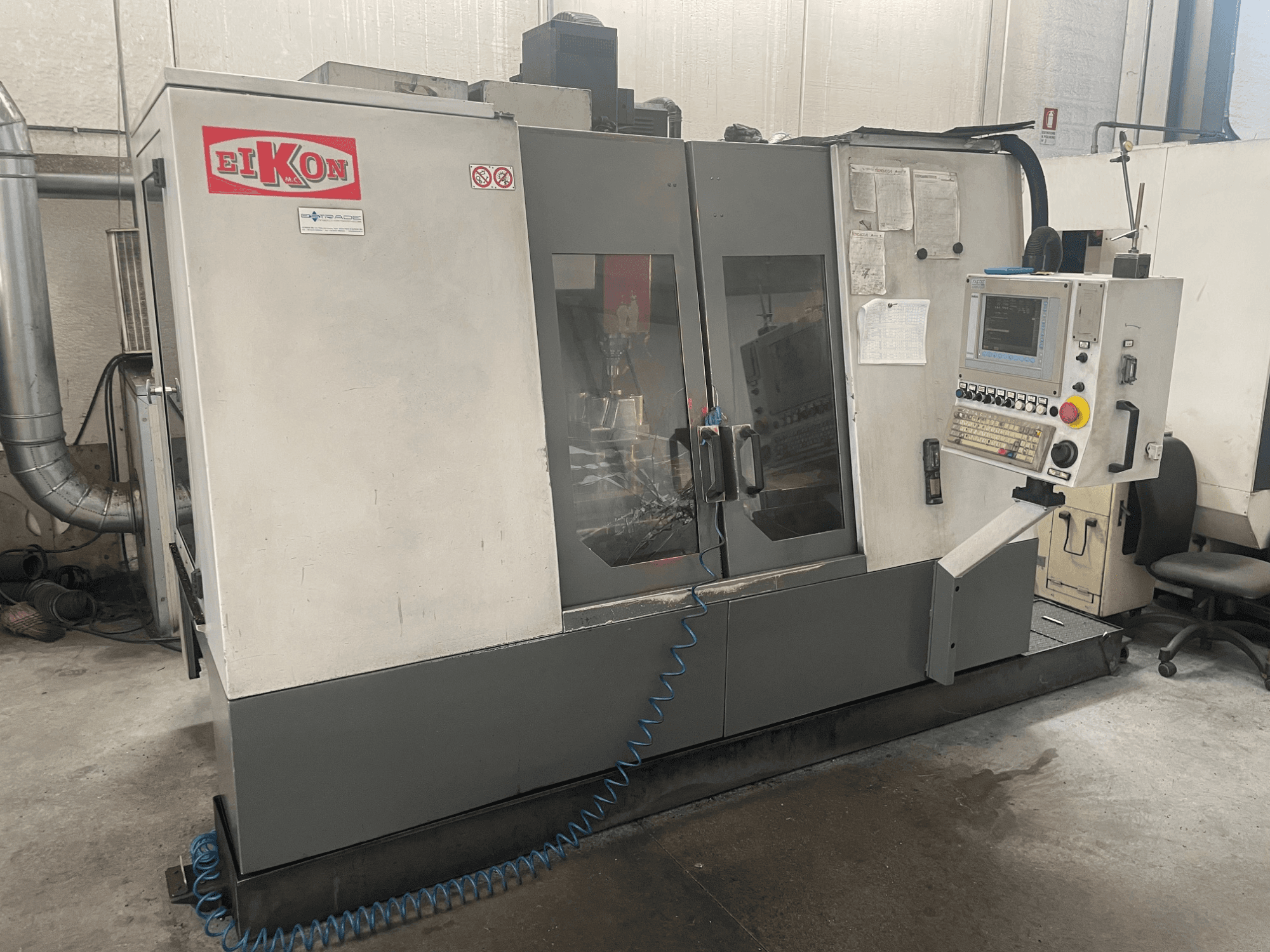 EIKON CNC stroj gledan sprijeda, s upravljačkom pločom, velikim staklenim vratima i plavim crijevom za zrak na podu.