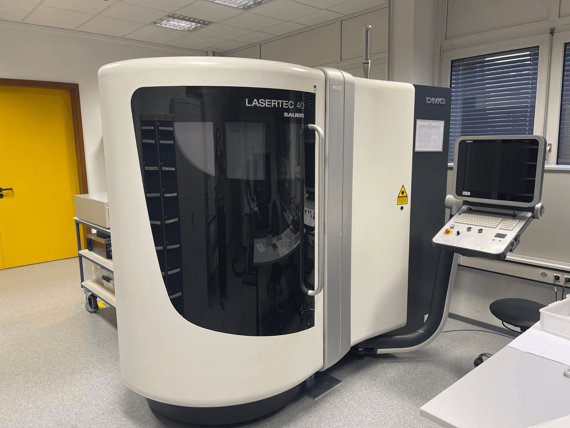 Bijela glodalica LASERTEC 40 tvrtke DMG MORI, pogled sprijeda, ima upravljačku ploču i ergonomski dizajn u dobro osvijetljenoj prostoriji.