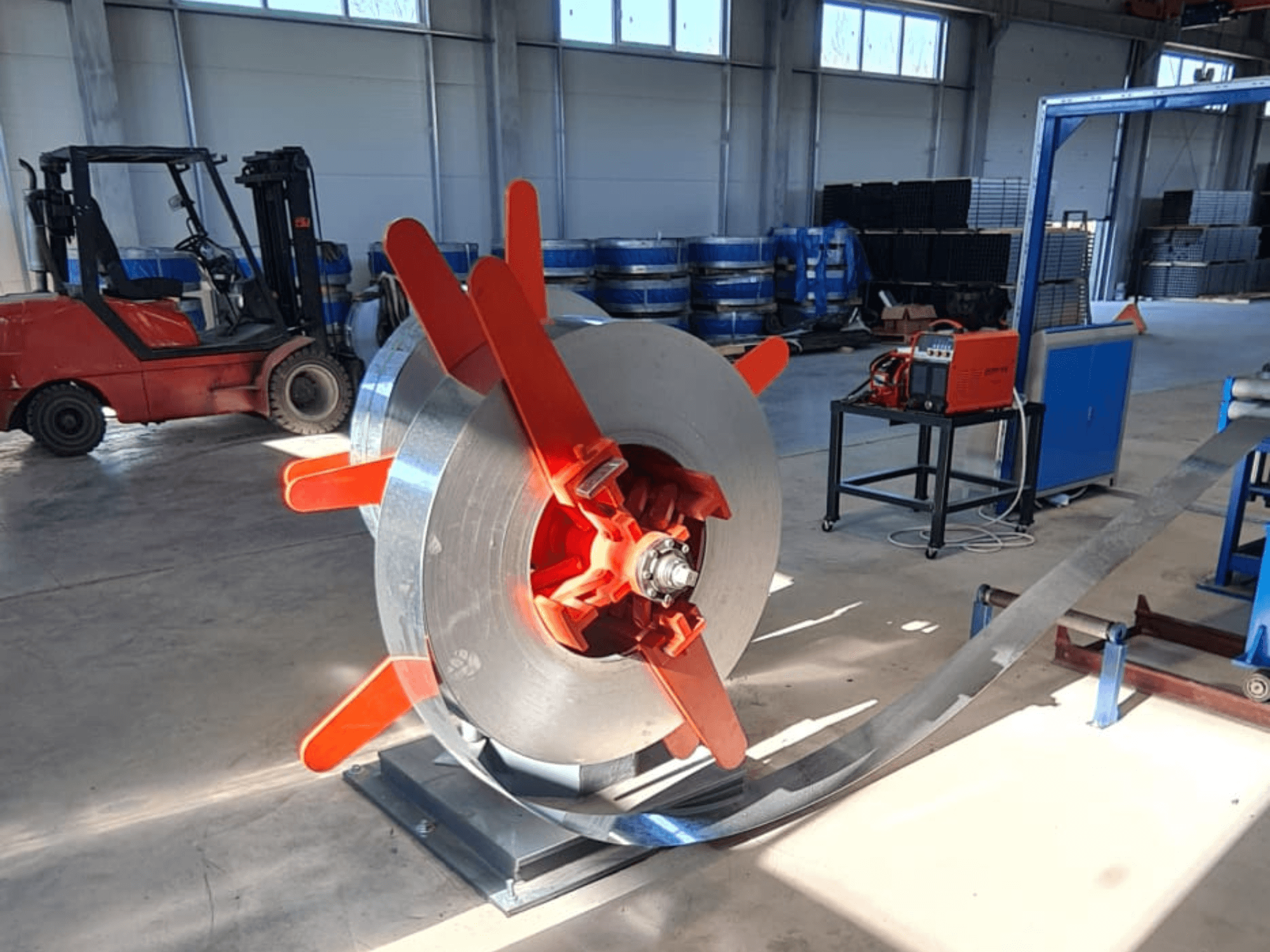 Metalna zavojnica s crvenim ručkama na stalku, gledano iz kuta. Industrijska pozadina sa strojevima i viličarom.