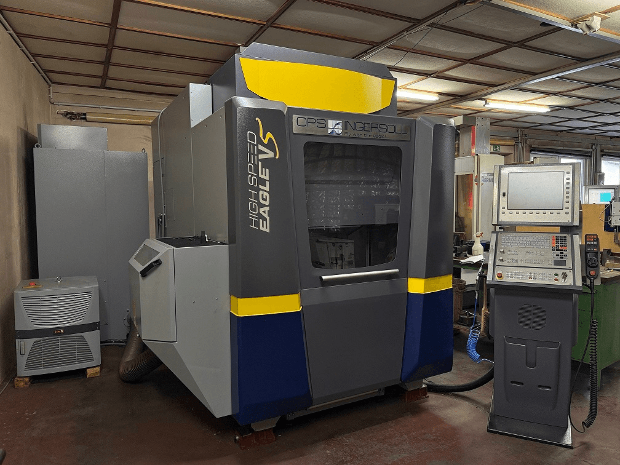 Stroj velike brzine Eagle V tvrtke OPS Ingersoll, bočni pogled, s upravljačkom pločom i susjednom opremom u industrijskom okruženju.