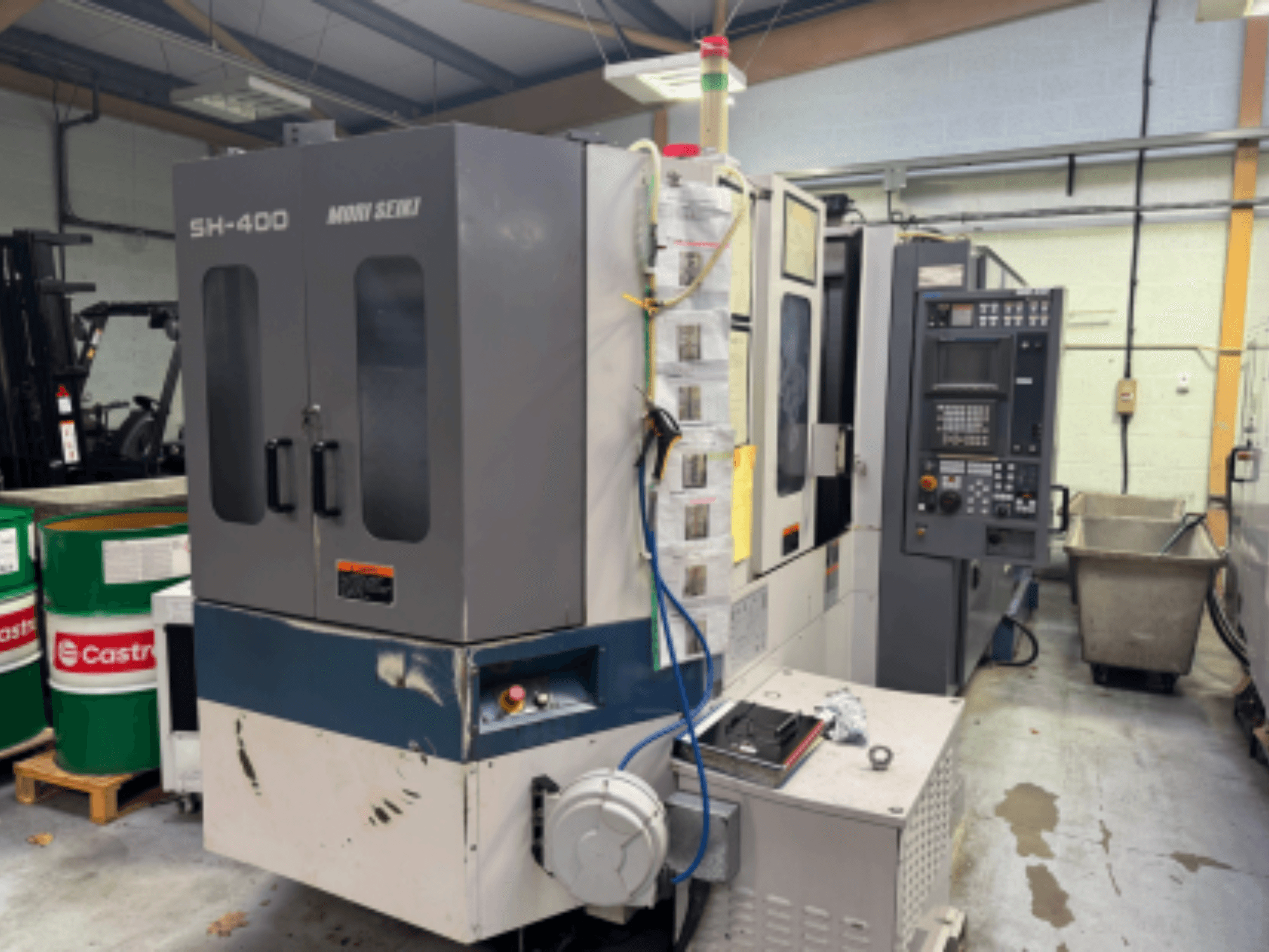 Moni Seiki SH-400 CNC stroj, bočni pogled koji prikazuje upravljačku ploču, vrata i okolno industrijsko okruženje.