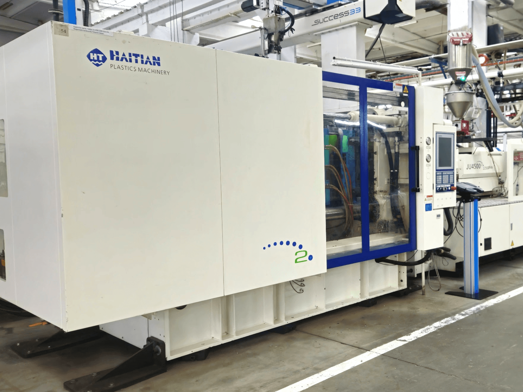 Model tvrtke Haitian Plastics Machinery, prednji pogled, prikazuje strojeve s upravljačkom pločom i vidljivim cijevima, u industrijskom okruženju.