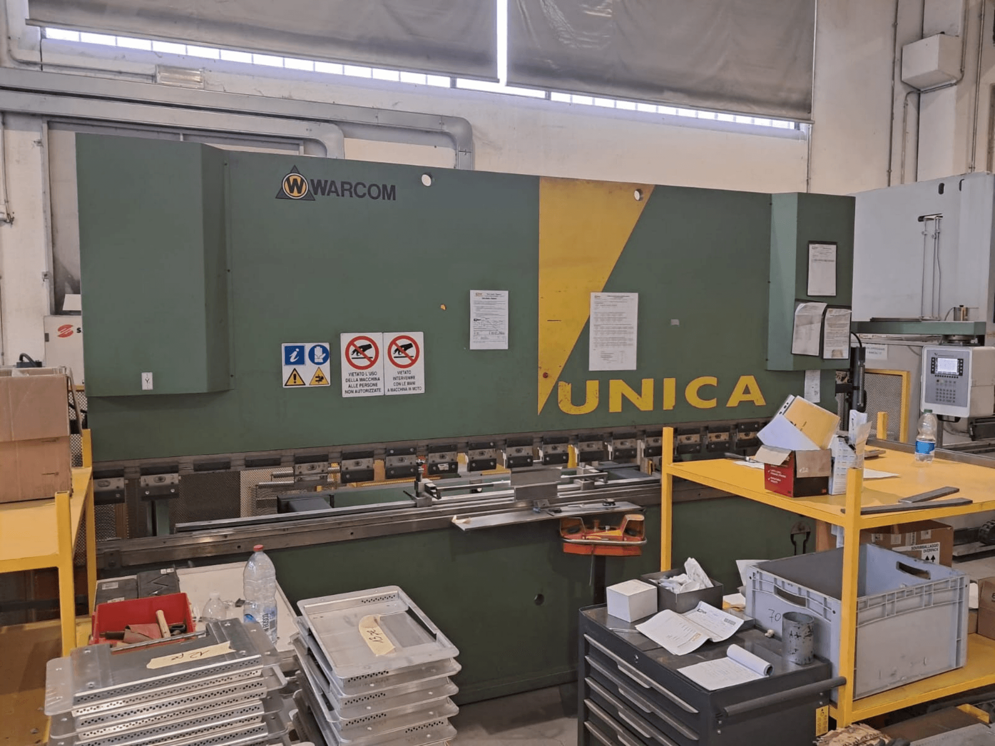 Hidraulična preša Warcom UNICA 100-40, prednji pogled, zeleno kućište sa žutim detaljima, vidljivi sigurnosni znakovi, alati i materijali na stolu.