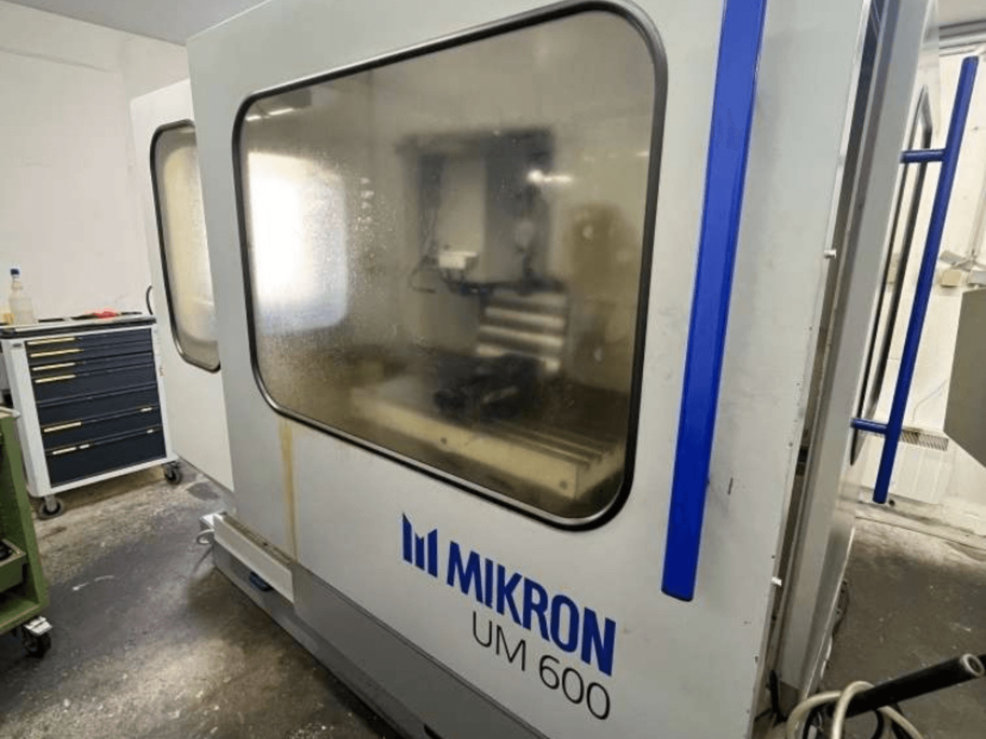 Univerzalni obradni centar MIKRON UM 600 TNC 410/426, bočni pogled, sa zamagljenim staklom i vidljivim skladištem alata u blizini.