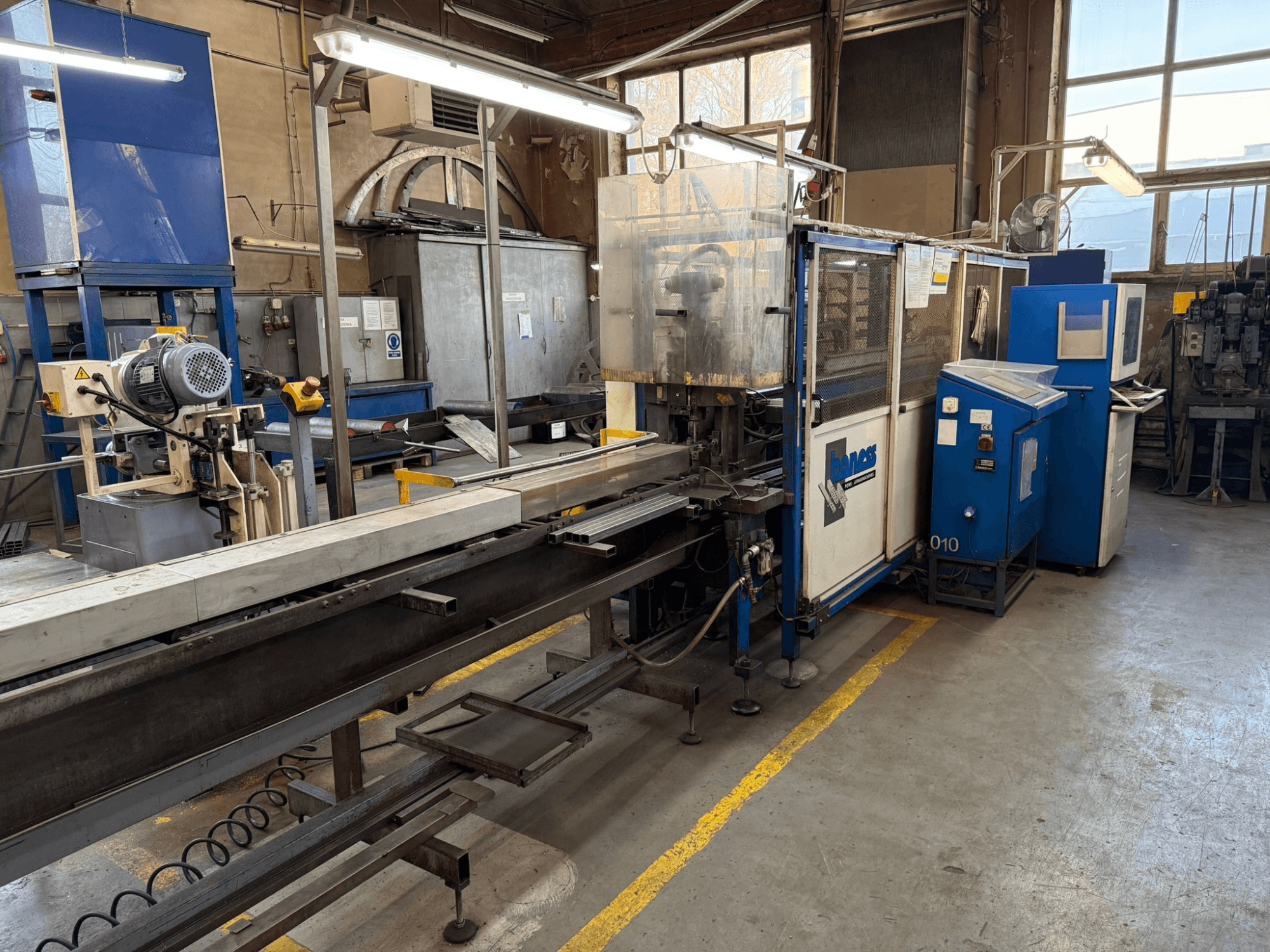 Industrijska radionica s plavim Haweas strojem, bočni pogled koji prikazuje transporter i opremu u velikom, dobro osvijetljenom prostoru.