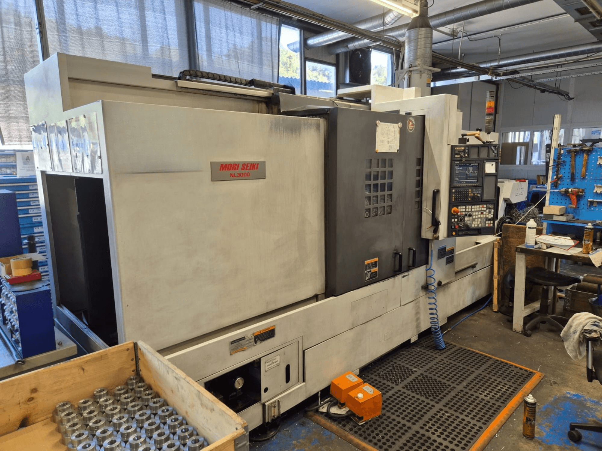 Mori Seiki NL3000 CNC tokarilica u radionici, bočni pogled s upravljačkom pločom, alatima i metalnim dijelovima u drvenoj kutiji.