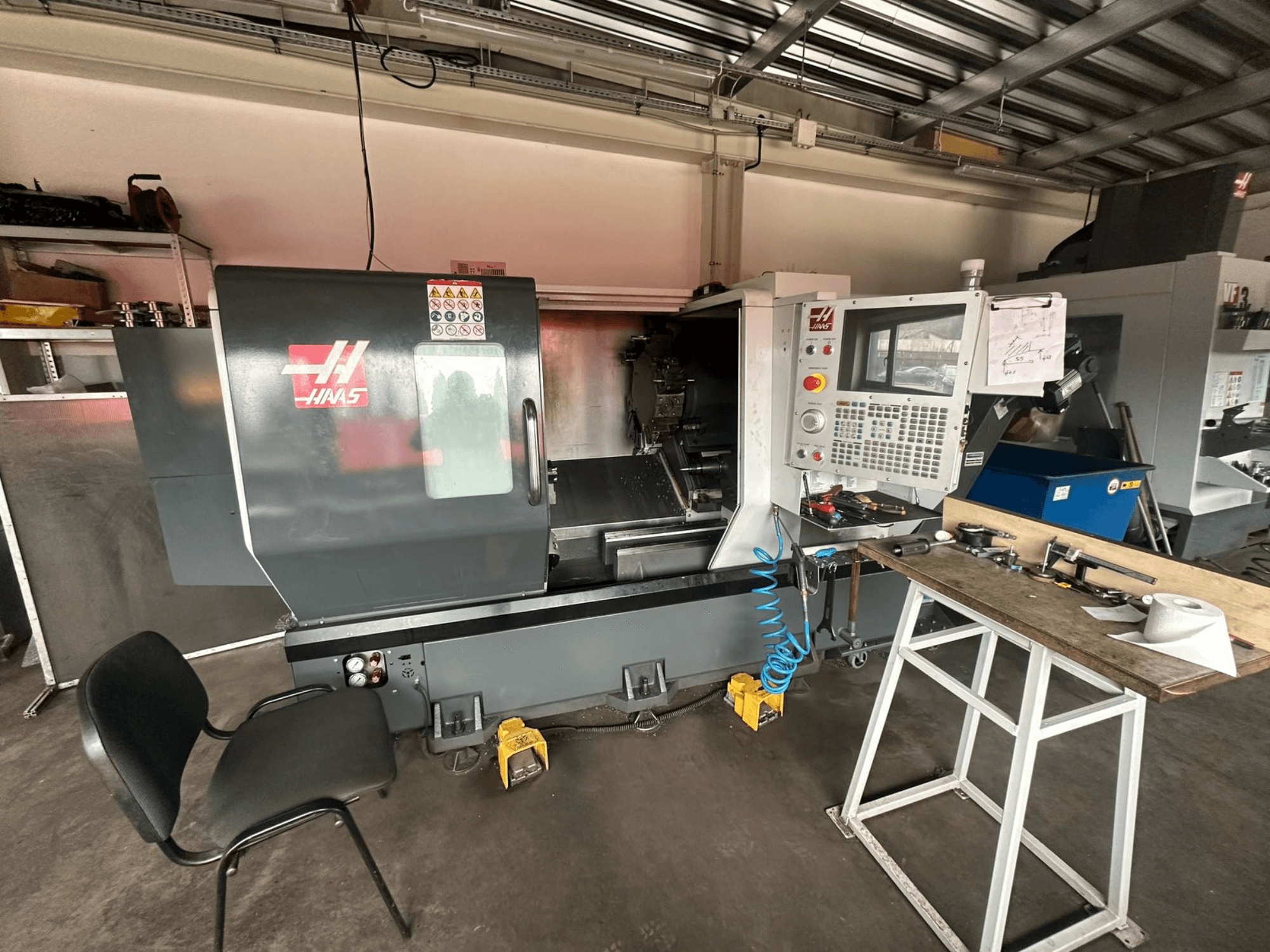 Haas CNC tokarilica (model ST-10) u radionici, bočni pogled s upravljačkim elementima, alatima na radnom stolu i stolicom u blizini.