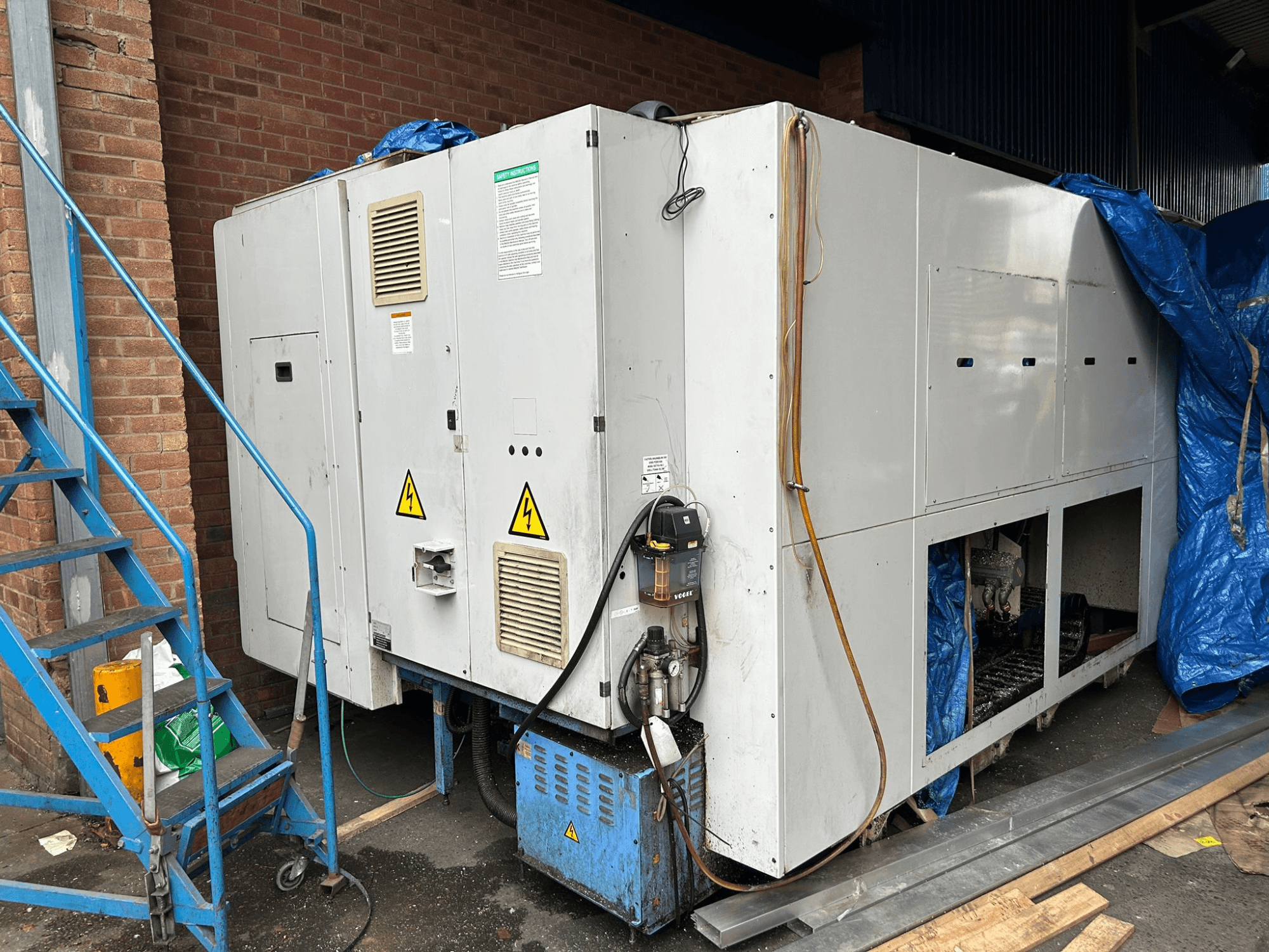 Bočni pogled na veliki industrijski hladnjak, nejasne marke i model, s prikazom električnih priključaka, znakova upozorenja i plave cerade.