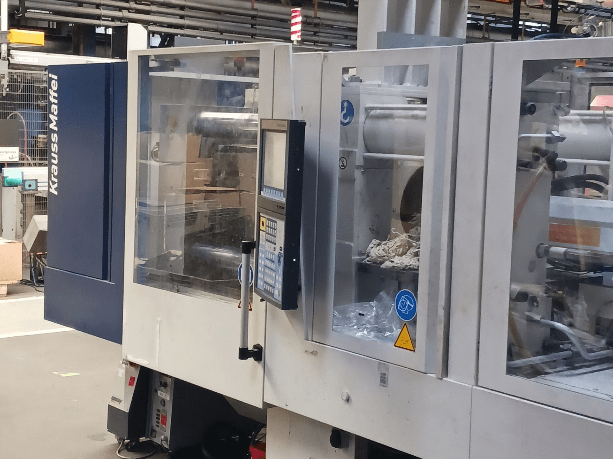 Stroj za brizganje plastike KraussMaffei, bočni pogled, s prikazom upravljačke ploče, strojeva i materijala za pakiranje u pozadini.