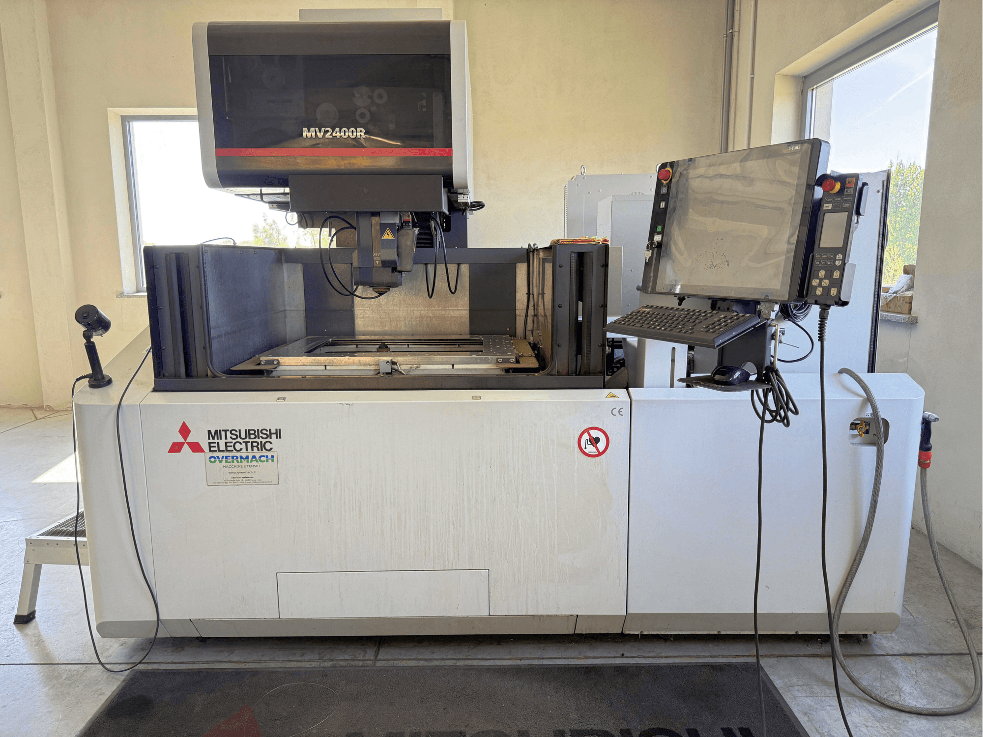 Stroj za žičanu erozijsku obradu Mitsubishi MV2400R u prednjem pogledu, s upravljačkom pločom, radnim prostorom i sigurnosnim znakovima.