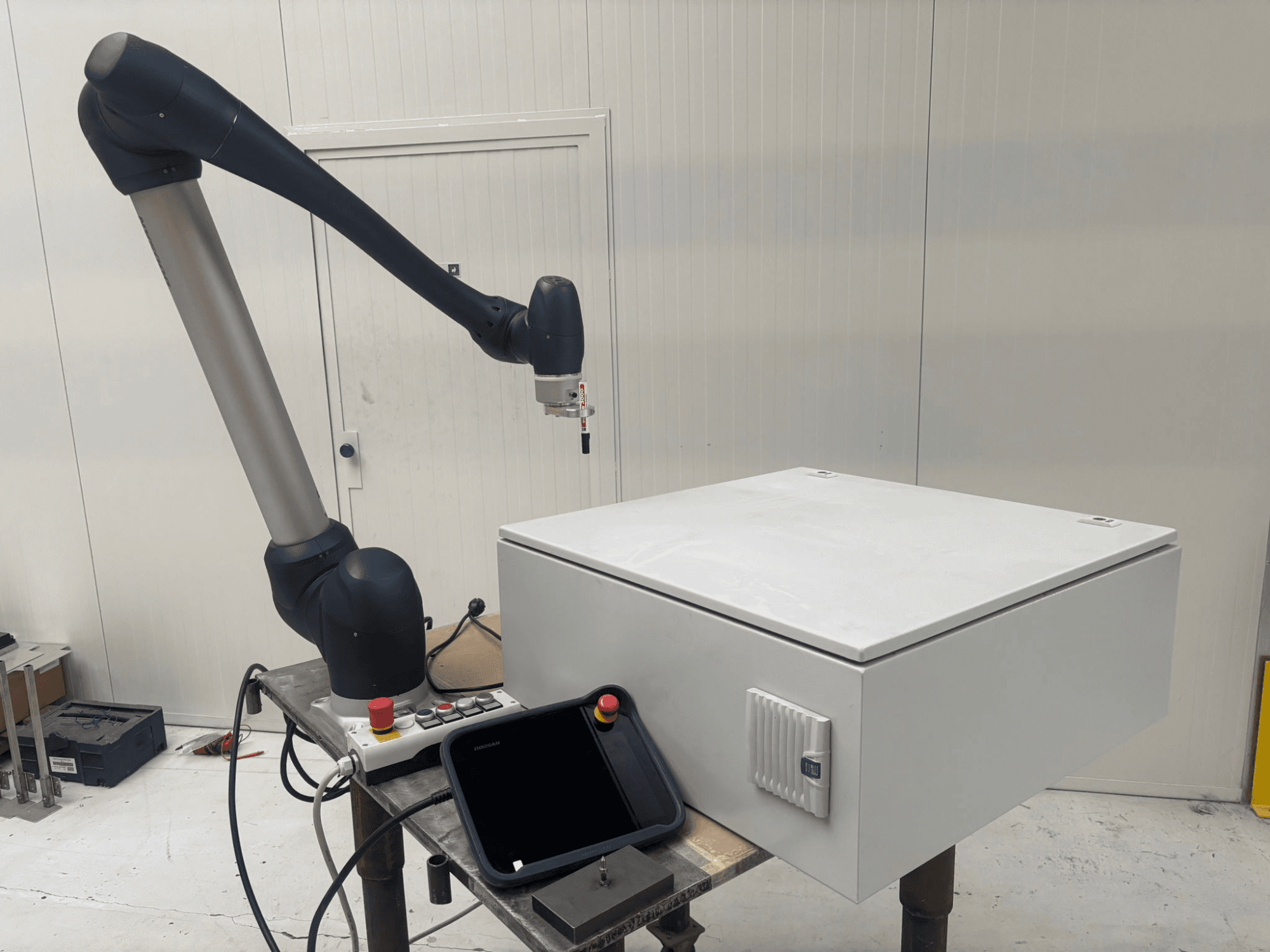 DOOSAN M0617 kolaborativni robot u bočnom pogledu, s robotskom rukom ispruženom iznad upravljačkog sučelja i bijelom kutijom na stolu.