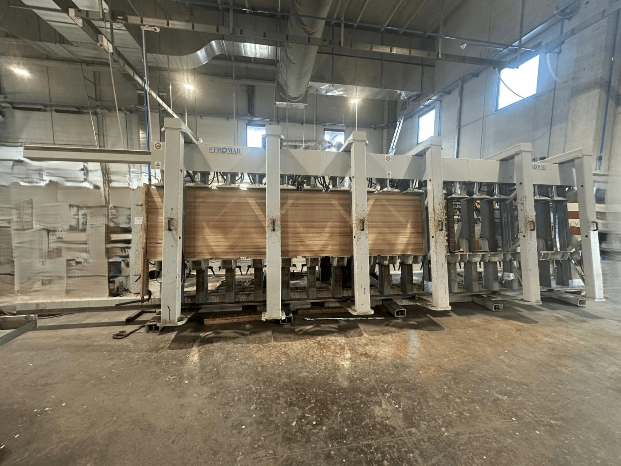 Industrijski STROMAB stroj u skladištu, pogled sprijeda, s drvenim pločama koje se obrađuju, s prikazom metalne konstrukcije i mehaničkih dijelova.